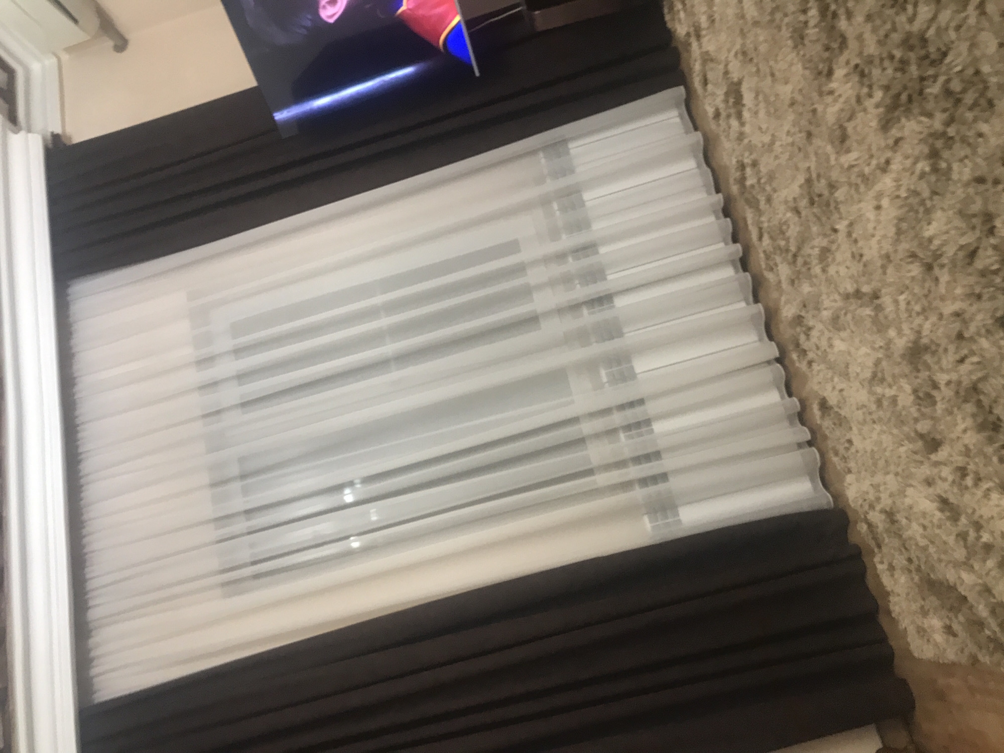 A low-angle, indoor shot shows a window covering with horizontal, white slats between brown fabric. Part of a TV and a beige shag rug are visible. There is no indication of the city of Yerevan, Armenia. There are no people, subjects, actions, or interactions visible.