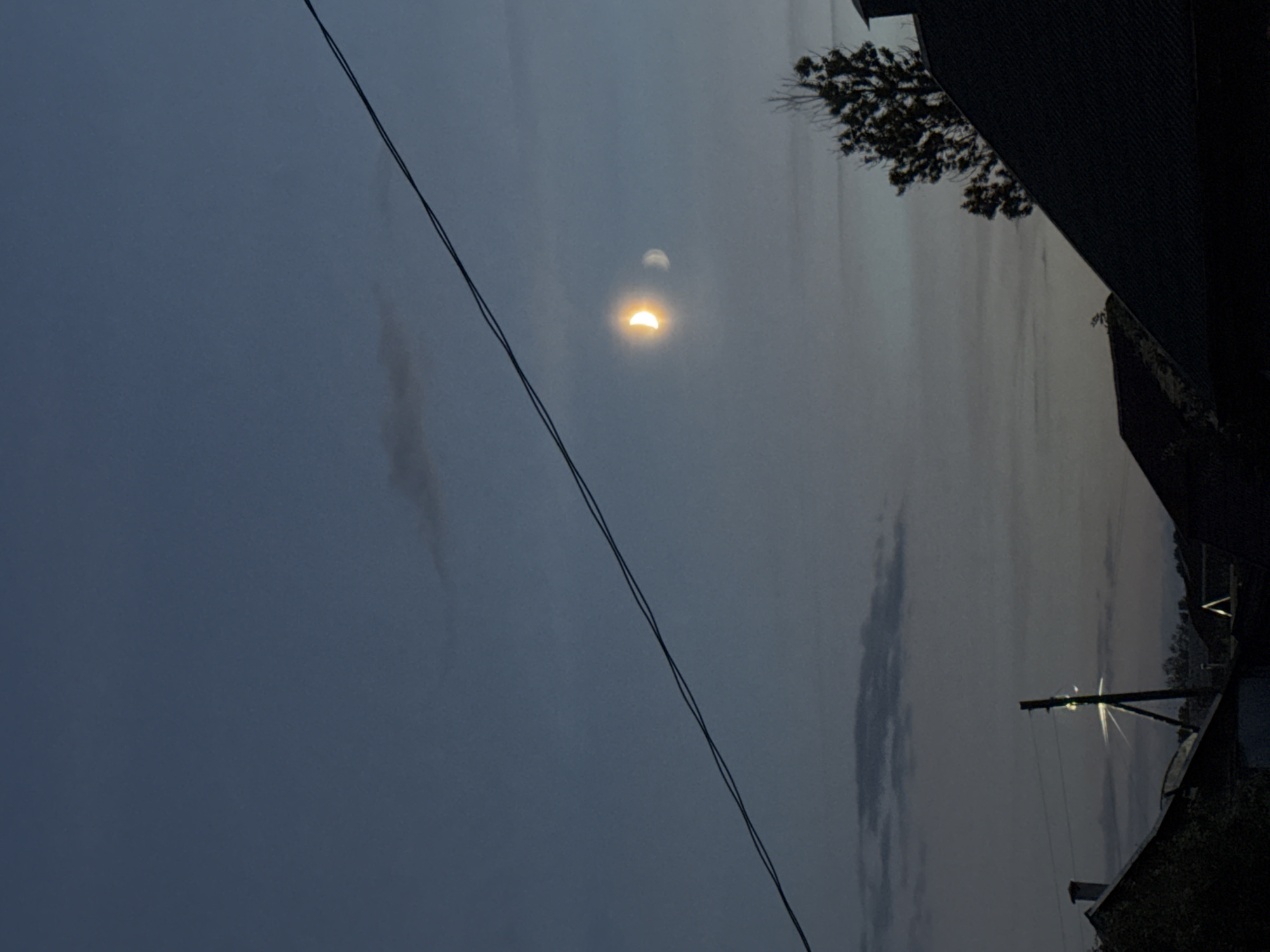 The image depicts a twilight sky over a residential area in Rzhev, Russia. A thin crescent moon, illuminated with a golden hue, is visible in the upper right quadrant of the sky, accompanied by a fainter, secondary moon-like image. Two dark power lines traverse the sky diagonally from the upper left to the lower right. In the foreground, the dark silhouettes of rooftops suggest houses. A tall utility pole with a bright light or reflection emanating from its top stands near the center of the frame. A silhouetted tree with sparse foliage is visible to the right of the pole, and a faint chimney stack is present on a rooftop in the upper left. The sky transitions from a deep blue at the top to a lighter, hazy gray near the horizon, with wisps of clouds scattered throughout.