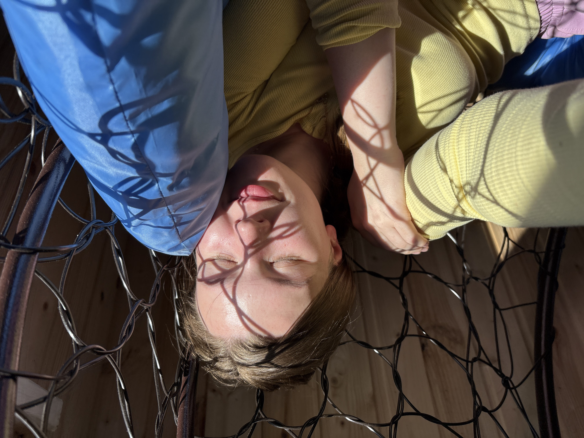 A young Caucasian female is depicted lying in a dark, woven metal hanging chair in Italmas, Russia. Her eyes are closed, and her facial expression is relaxed, indicating rest or sleep, with a slight smile present on her lips. She is wearing a light yellow, ribbed long-sleeved top, and a portion of light purple clothing is visible at the bottom left. Her head rests against a bright blue fabric cushion or pillow, which also supports her right arm. Strong sunlight illuminates the scene from the upper left, casting intricate, sharp shadows of the metal chair's grid pattern across her face, right arm, and the blue cushion. The background consists of light-colored wooden planks, visible through the gaps in the metal structure, suggesting an indoor or sheltered outdoor setting.