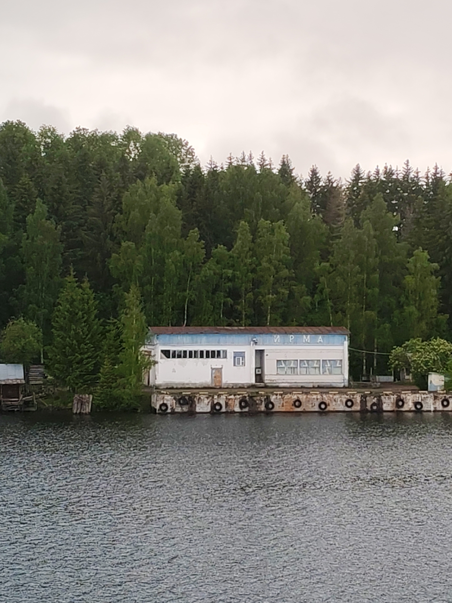 The image captures a weathered, single-story building located on a concrete pier. The building, possibly an old commercial or industrial structure, displays the Cyrillic word "ИРМА" on its facade. The pier is lined with old tires along the edge, presumably as bumpers. Dense forest forms a backdrop to the building and pier. In the foreground, calm water fills the frame. The sky is overcast. This is a scene of the Раменье area in Russia.