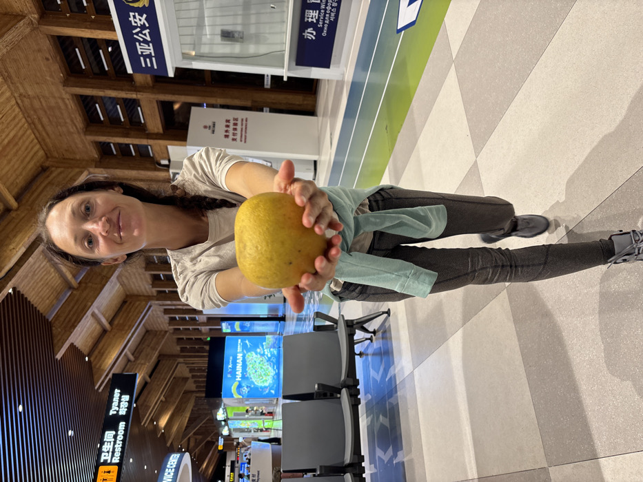 The image captures a female individual holding a large yellow citrus fruit within Sanya Phoenix International Airport (SYX) in Tianya District, Sanya, Hainan, China. The woman, positioned centrally and facing the camera, has dark hair, wears a light short-sleeved top, dark grey patterned leggings, and light grey athletic shoes, with a light blue shirt tied around her waist. She extends both hands forward, presenting the fruit towards the viewer, and exhibits a slight smile.
The environment is a modern airport terminal featuring a distinctive wooden slatted ceiling and a light-colored tiled floor composed of grey and beige squares. Bright artificial lighting illuminates the scene. In the background to the left, airport seating (dark grey, bench-style) is visible, along with a black "Restroom" sign displaying text in Chinese, English, and Korean. Further back, illuminated display boards include one with "HAINAN" and an underwater image. To the right in the background, white service kiosks are present, one featuring a blue sign with "三亚公安" (Sanya Public Security) in Chinese and partially visible English text. Another sign for "HILTON" is also discernible.