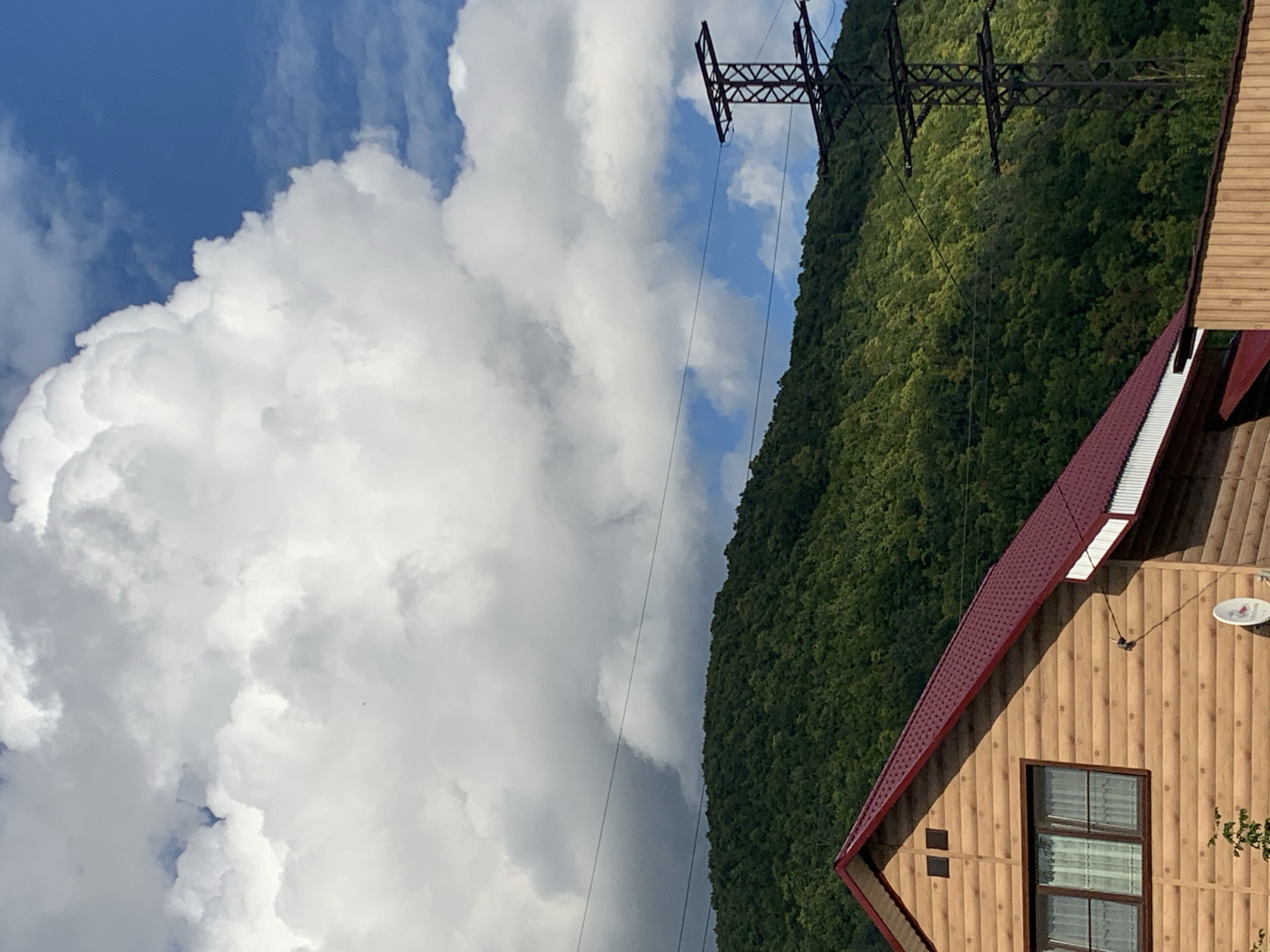 The image captures a view from a residential area towards a densely forested mountainside under a partly cloudy sky. In the foreground, the side of a wooden house with a dark red metal roof is visible. The house features a window with white curtains and a satellite dish mounted on the wall. A wooden fence runs along the bottom of the image. In the middle ground, a large pylon for high-voltage power lines stands against the backdrop of the green mountain, with several power lines crisscrossing the frame. The mountain is covered in lush, dark green trees, suggesting a temperate climate. The sky is a vibrant blue, interspersed with large, puffy white clouds. The scene appears to be located in the city of Asha, Russia, a region known for its mountainous terrain and forests.