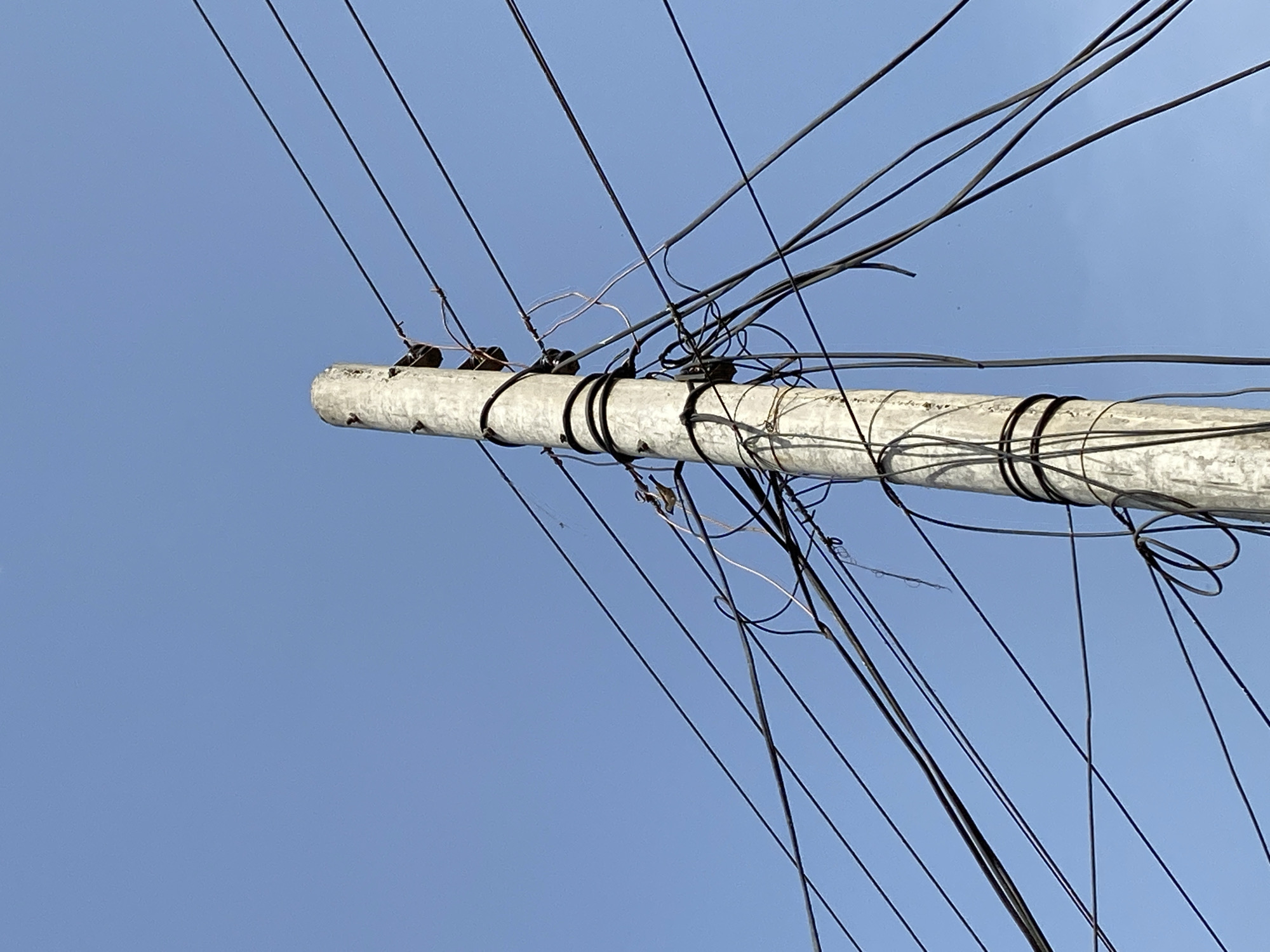 The image shows a utility pole against a clear blue sky in Chak 166 GB, Pakistan. The pole is made of concrete and has a rough, weathered surface. Numerous black electrical wires are attached to the pole with metal brackets and bindings. The wires run in various directions, creating a cluttered appearance. No people or other objects are visible in the image.