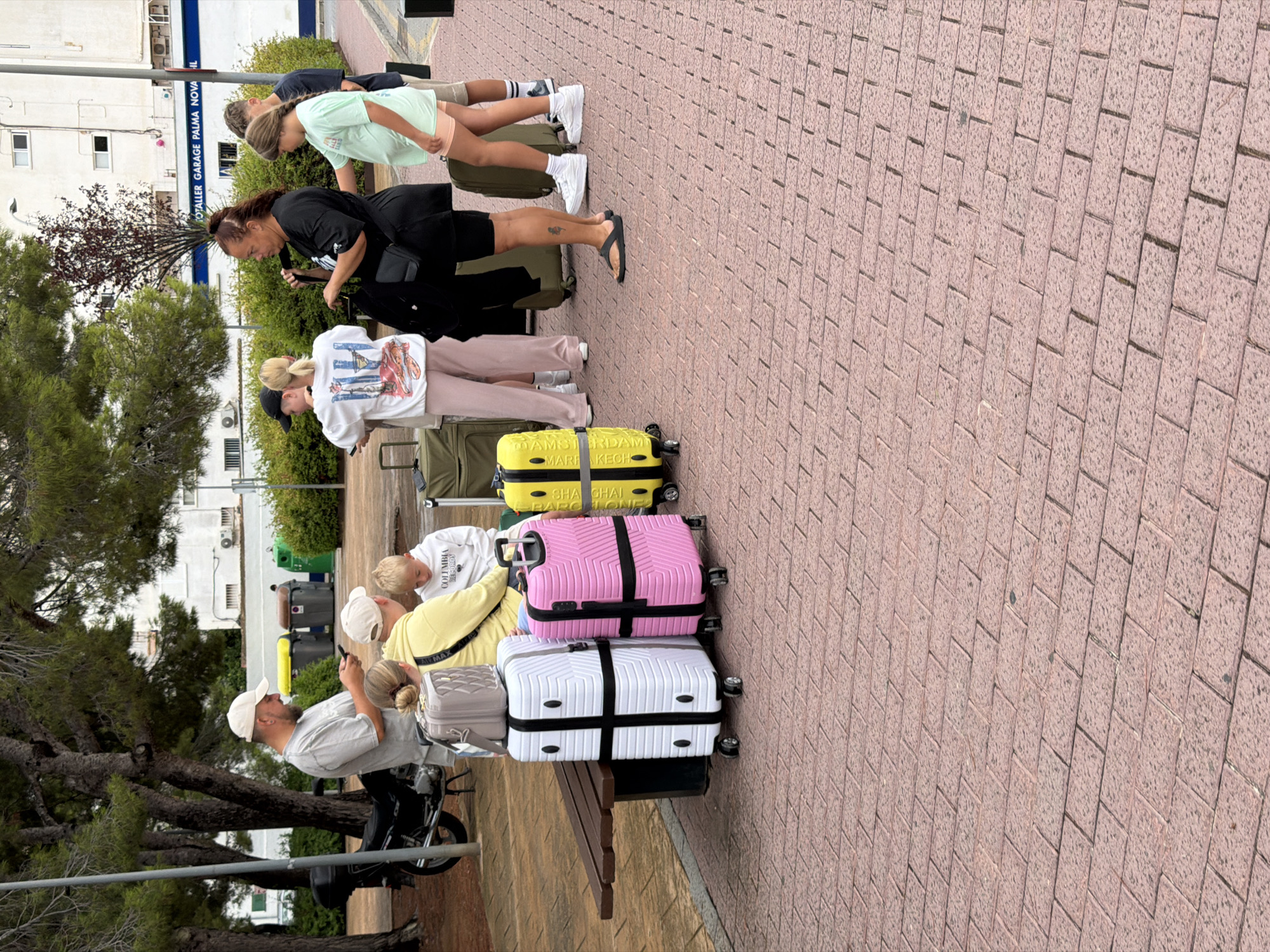 In the city of Calvià, Spain, a group of people are gathered outdoors on a brick-paved walkway, with a backdrop of trees, buildings, and parked vehicles. Two adults, one male wearing a light-colored baseball cap and t-shirt, and another female with dark hair pulled back, are standing. A young boy in a white hoodie and a girl in a light green t-shirt and shorts are also present. Several suitcases are arranged around the group, including a pink and a white hard-shell suitcase, a green soft-sided suitcase, and a smaller grey carry-on. A motorcycle is parked to the left of the frame. In the background, buildings with windows and air conditioning units are visible, along with a sign that reads "TOTALER GARAGE PALMA NOVA IL". The overall scene suggests a moment of waiting or preparation for travel.