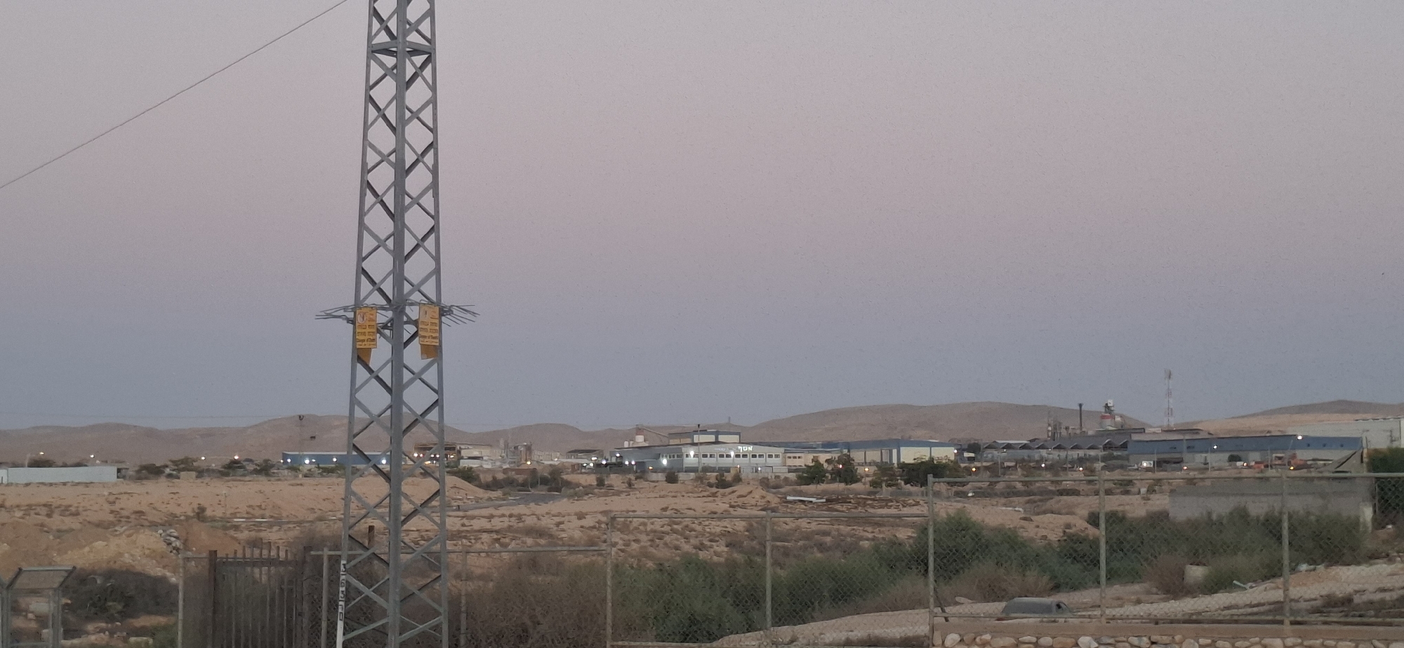 The image captures a wide-angle view of an industrial area in Yerocham, Israel, likely at dusk given the soft, diffused light and the pale sky. In the foreground, a tall, grey lattice metal pylon dominates the left side of the frame, with two yellow signs attached to it. A chain-link fence runs across the lower part of the image.
Beyond the fence, the landscape is arid and desert-like, with scattered scrub vegetation. A cluster of industrial buildings forms the mid-ground, characterized by their low-rise structures, some with blue roofs, and various chimneys and towers. Some lights are visible in the buildings, indicating ongoing activity. The background consists of rolling, barren hills under a hazy, pale sky. The overall impression is one of a functional, industrial zone within a desert environment. There are no people or animals visible in the image.