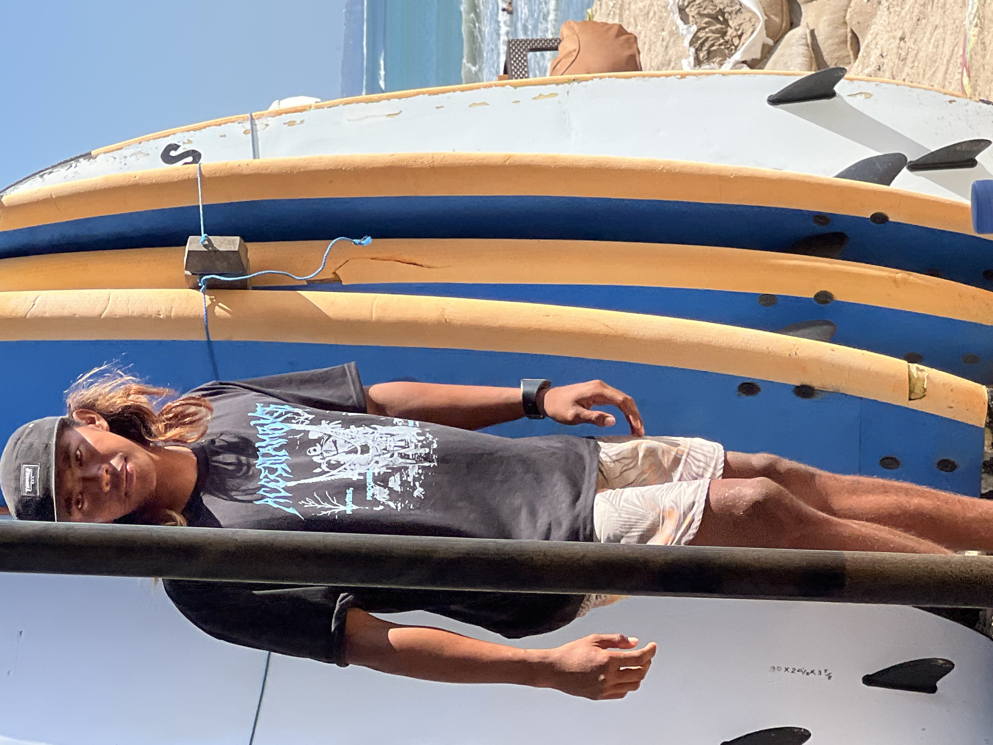 A young person with long, wavy brown hair and wearing a black t-shirt with a white graphic design and a black baseball cap is lying down on a stack of surfboards. They are looking towards the camera with a relaxed expression. The surfboards are stacked horizontally, with alternating blue and orange sections. The person is wearing patterned shorts. A black smartwatch is visible on their left wrist. The scene appears to be outdoors, with the ocean and beach visible in the background. The lighting suggests it is daytime, likely sunny. The location is identified as Kuta, Indonesia. The surfboards have fins attached. Some text is visible on the bottom surfboard, "30 X 24 1/2 X 3 1/8".