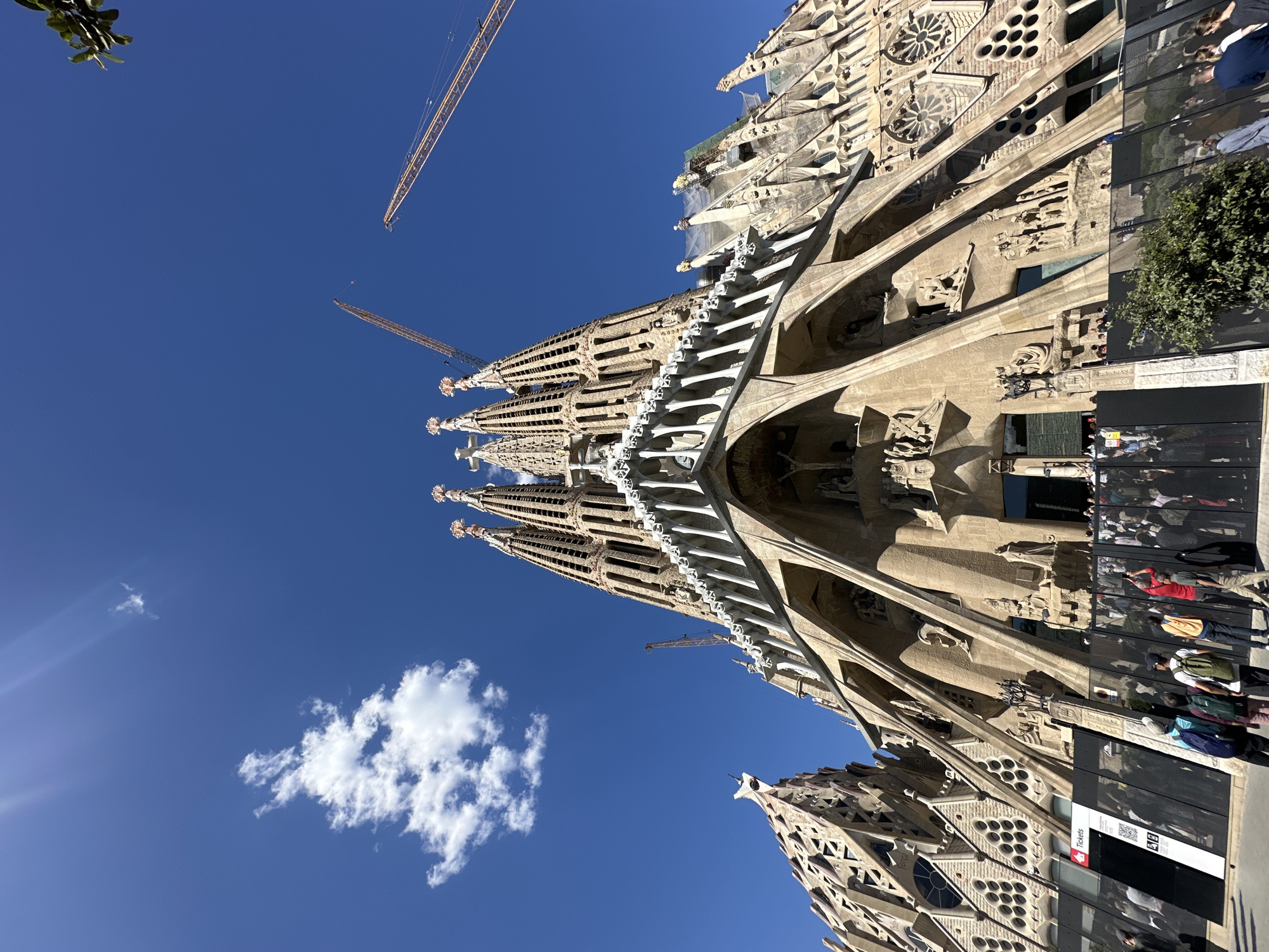 The image captures an upward, angled view of the Sagrada Familia basilica in Barcelona, Spain, set against a clear, deep blue sky with a single white cloud. Construction cranes are visible extending above the basilica's spires, indicating ongoing building work. The basilica's intricate stone facade is detailed, and a group of people are standing near the entrance. There are reflective surfaces visible on the right side of the image with people and signs.
