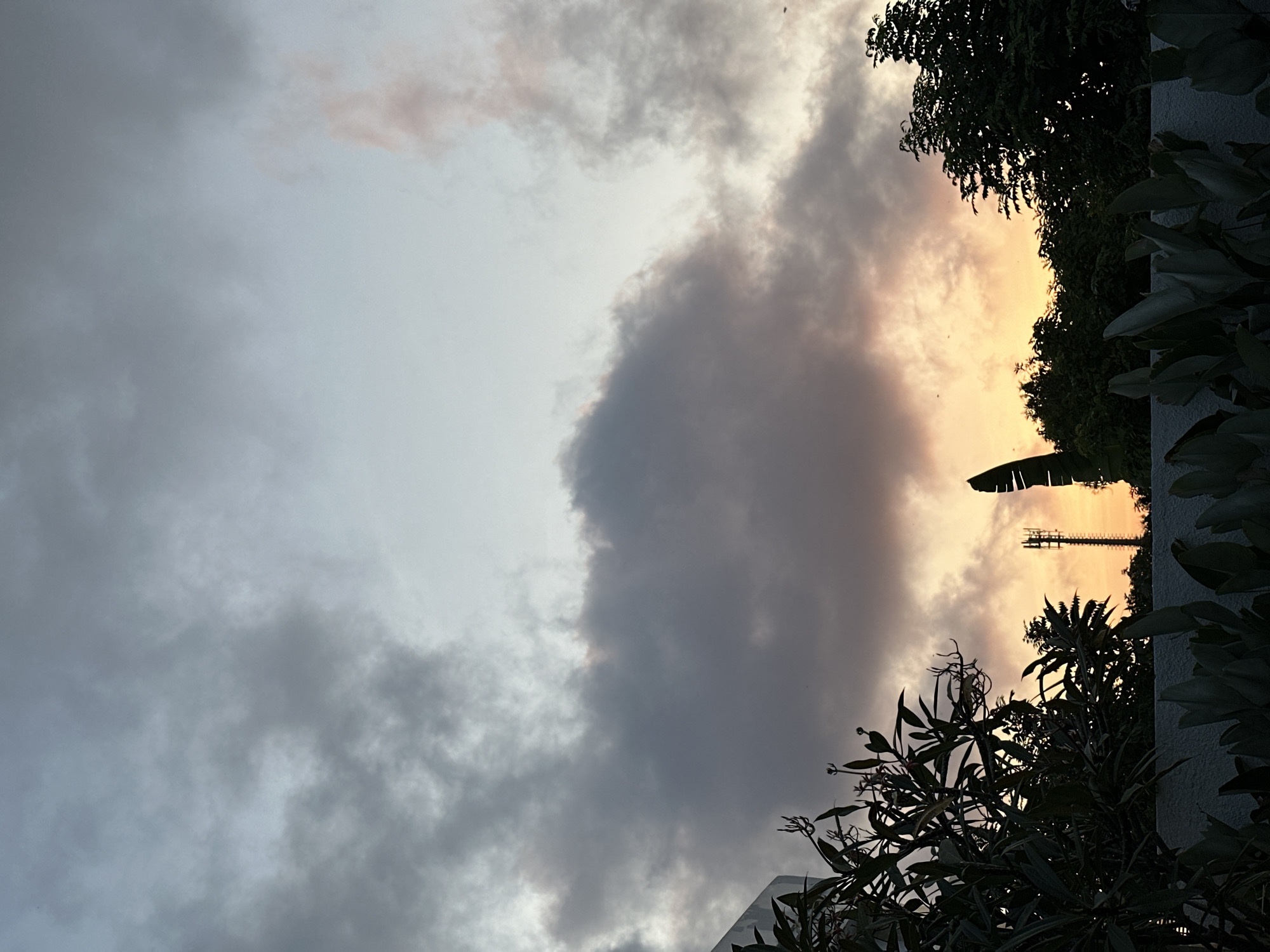 This photograph captures a sunset scene in Kuta Selatan, Indonesia. The sky is filled with dramatic clouds, ranging in color from dark gray to soft pink and bright orange, indicative of the twilight hour. In the mid-ground, a cellular tower stands tall, silhouetted against the colorful sky, positioned just above a line of dense, dark green trees. In the foreground, the tops of lush, tropical plants and foliage are visible, along with a white wall. The overall impression is of a serene, natural landscape illuminated by the fading light of the sun.