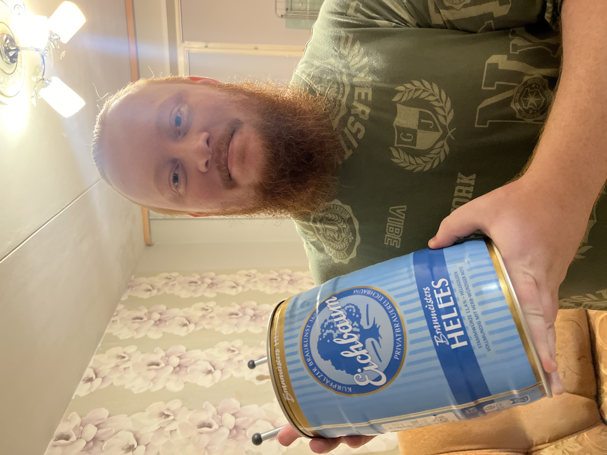 A man with a prominent red beard and short reddish hair is captured indoors, looking directly at the camera with a neutral expression. He is holding a large blue and gold mini-keg of German "Eichbaum" beer. The man wears a dark green t-shirt adorned with faded light-colored graphics, including partial words like "SITY," "VIBE," "ORK," and a crest featuring the letters "G J" and a laurel wreath, suggesting a university or sports theme.
The "Eichbaum" mini-keg prominently displays the brand's tree logo and various texts: "KURPFALZER BRAUKUNST 1629," "PRIVATBRAUEREI EICHBAUM," and "Braumeisters HELLES." Additional details on the keg indicate "STAMMWÜRZE 11,4% - HELLGOLDEN" and "VOLLMUNDIG MIT HERB-WÜRZIGER NOTE." Two black spigots or handles are visible on the top of the keg.
The indoor setting, likely a room in Chaikovsky, Russia, features a wall with light-colored floral wallpaper, displaying rows of delicate light pink or purple blossoms on a pale background. Above the man, a white ceiling fan with three illuminated light fixtures provides bright artificial light. A glimpse of light brown upholstered furniture is visible in the lower right corner, indicating a relaxed domestic environment. The image is brightly lit by the artificial lights, making the time of day indeterminate.