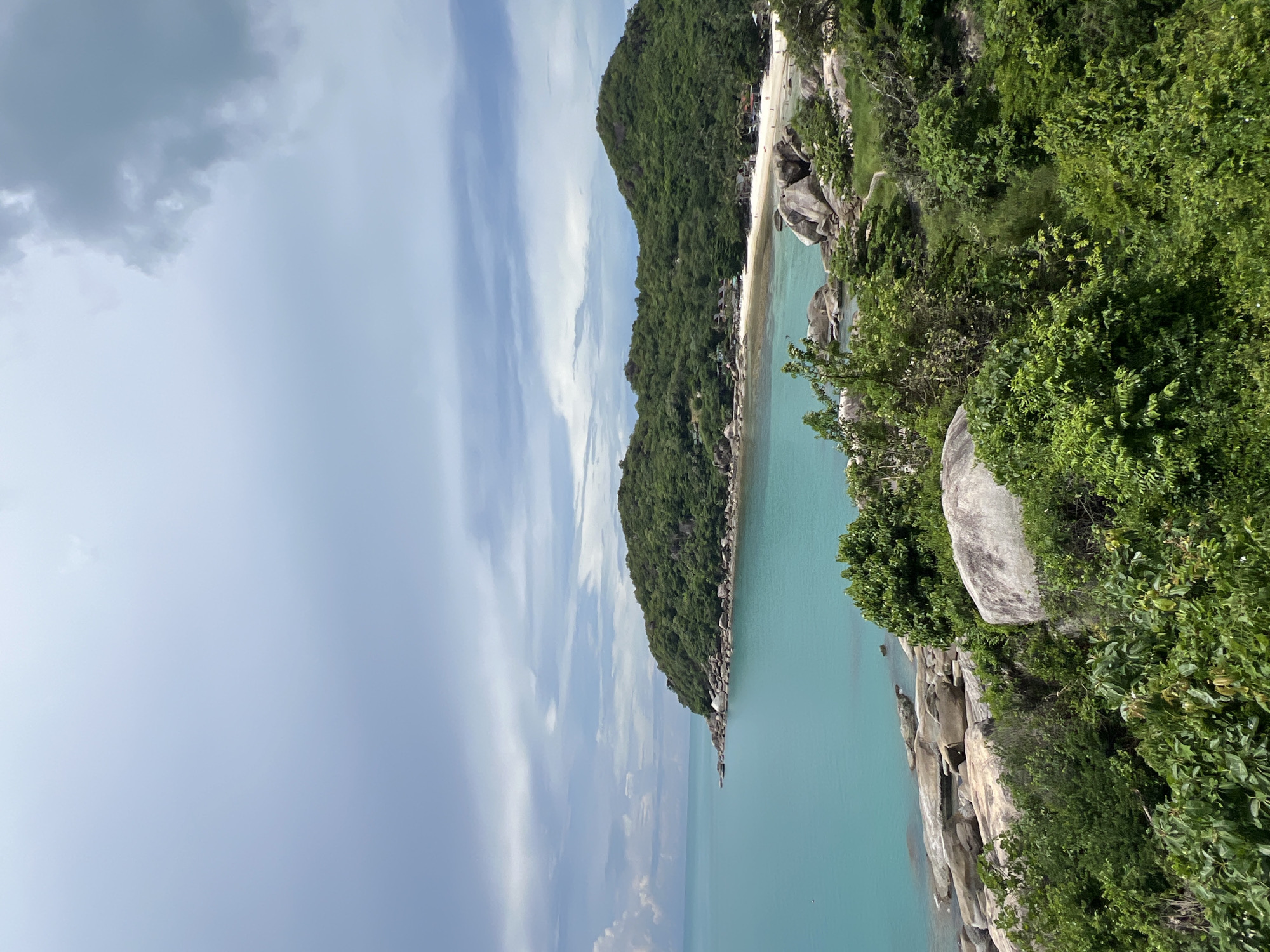 The image shows a scenic view of a tropical coastline with lush green hills meeting a turquoise sea under a cloudy sky. In the foreground, dense green foliage and large boulders are visible. The middle ground features a sandy beach with scattered buildings and more large rocks along the shoreline. The background consists of rolling, tree-covered hills extending out into the ocean. The water appears calm with a slight ripple. The sky is filled with a mix of white and gray clouds. The location is identified as Baan Chaweng Noi, Thailand. There are no people or identifiable actions depicted in the scene.