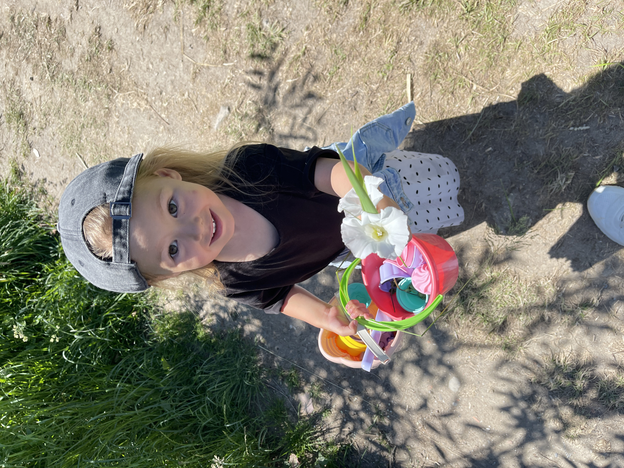 A young child wearing a grey baseball cap and a black t-shirt smiles at the camera, holding a red bucket filled with colorful plastic toys and two white flowers. The child is also wearing a denim jacket over a white dress with black polka dots. They are standing outdoors on a sunny day, with grass and dirt ground visible. The shadow of a tree indicates it's likely daytime. The overall impression is one of childhood joy and outdoor play.