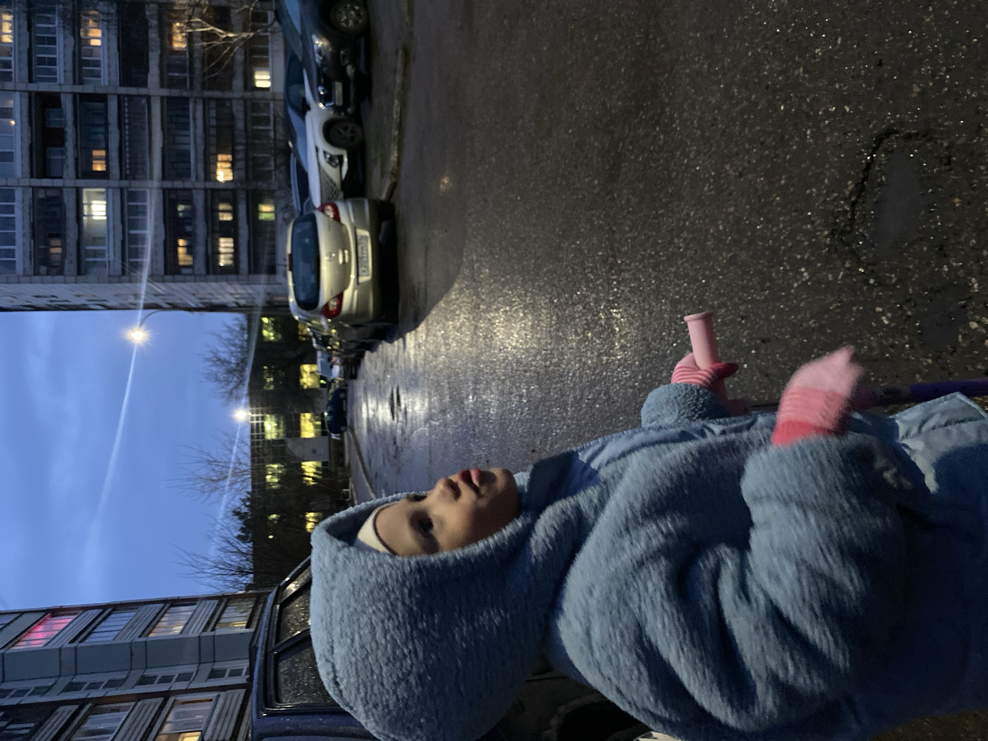 This image, taken in Chaikovsky, Russia, captures a young child during dusk or early night on a wet urban street.
The primary subject is a young child, likely a toddler or preschooler, positioned in the foreground on the right side of the frame. They are wearing a fluffy, light blue hooded coat and a white cap or bonnet. The child's face is visible, looking upwards and slightly to the left, with a neutral expression and slightly parted lips. Pink and white striped gloves or mittens cover their hands, with one hand holding a pink, cylindrical object, possibly a toy handle.
The setting is an outdoor residential area, likely a street or courtyard flanked by multi-story apartment buildings. On the left side of the image, a large, grey apartment building stands tall, characterized by numerous windows, many of which are illuminated with warm and cool lights, suggesting habitation. Bare tree branches are visible against the twilight sky.
The ground is a dark, wet asphalt surface, reflecting the ambient lights from the buildings and a bright street lamp visible on the left. This wetness suggests recent rain or melted snow. Several cars are parked along the side of the street. One silver hatchback, partially visible, has a license plate that clearly shows "У293МТ". Other darker vehicles are also parked nearby.
The sky above is a deep blue twilight, with a distinct white contrail stretching across it. A street lamp on the left provides additional illumination to the scene. The overall impression is one of a quiet, everyday evening in a residential district during cooler weather, with the child seemingly observing their surroundings.