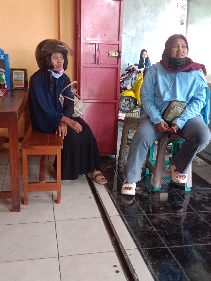 Two women are sitting on chairs. The woman on the left wears a black helmet, dress, and sandals and carries a patterned purse. The woman on the right is in a light blue top, jeans, and white platform sandals. She wears a dark headscarf and carries a small bag. A yellow scooter is visible in the background, behind a third person in a headscarf. The scene appears to be indoors, near a doorway and tiled flooring.