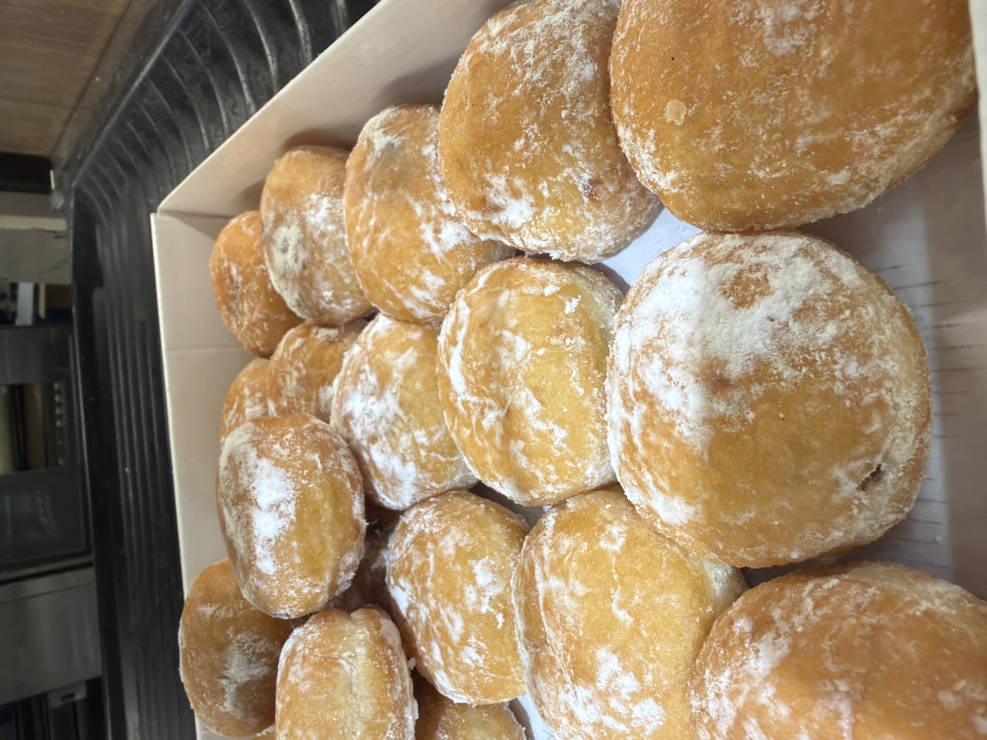 Approximately twenty golden-brown, round pastries, consistent with donuts or *ponichki*, are densely arranged within a white, open cardboard box. Each pastry is uniformly coated with white powdered sugar. The pastries exhibit a smooth, slightly irregular texture indicative of fried dough. In the background, a dark, corrugated surface, likely a commercial baking tray or countertop, is visible behind the box. Further back, indistinct dark objects, possibly kitchen equipment, and a light-colored wooden surface are present. The scene is set in a commercial bakery or kitchen environment in Sofia, Bulgaria. No individuals are present, and no actions are depicted.