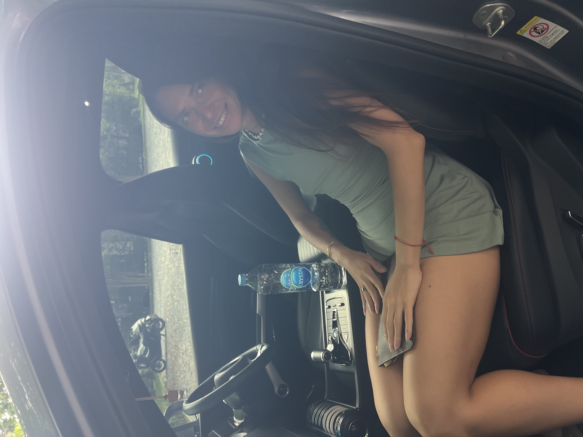 A smiling young woman with long dark hair is captured inside a car, looking towards the camera. She is wearing a light green sleeveless dress or romper and a beaded necklace. A red string bracelet is visible on her left wrist, and a gold bracelet on her right. Her posture suggests she is leaning or kneeling in the car's interior, possibly near the passenger seat or center console. She holds a gray cloth or rag in her right hand, near her thigh.
A clear plastic bottle of "CRYSTAL" brand water is placed on the dashboard or center console to her left. Parts of the car's interior, including the steering wheel, dashboard, and a gear shifter on the center console, are visible. Through the car windows, an outdoor environment with greenery and what appears to be a building or wall can be seen. A motorcycle is partially visible outside the car's window.
Inside the door jamb, a yellow warning sticker with "Attention" at the top is present, featuring symbols that likely pertain to car safety, though the smaller text is illegible. The woman's expression is cheerful. The image appears to be taken during daylight hours. The overall scene is set in a car, with the provided context indicating the location is บ้านบางรักษ์, Thailand.