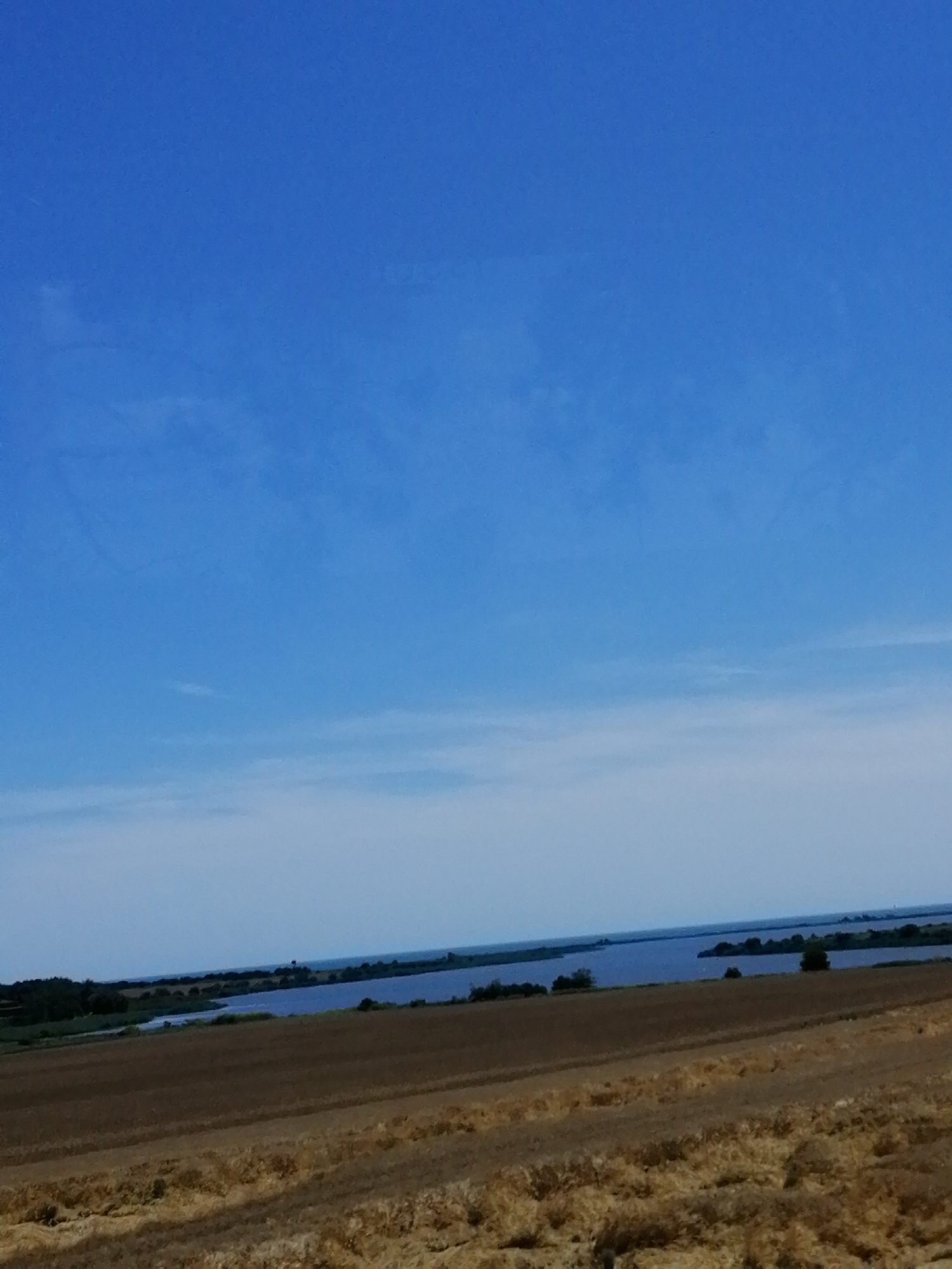 The image depicts a landscape in Durankulak, Bulgaria, under a clear blue sky with faint wispy clouds. In the foreground, a harvested field of dry, golden stubble stretches across the frame, showing the texture of the tilled earth and cut stalks. Beyond the field, a body of water, likely Durankulak Lake, is visible, reflecting the sky and appearing as a strip of deep blue. The far shore of the lake is lined with a dark green, dense line of trees and vegetation, creating a stark contrast with the agricultural land. The horizon line is defined by the meeting of the lake and the distant treeline. The overall impression is one of rural tranquility and the vastness of the open sky.
