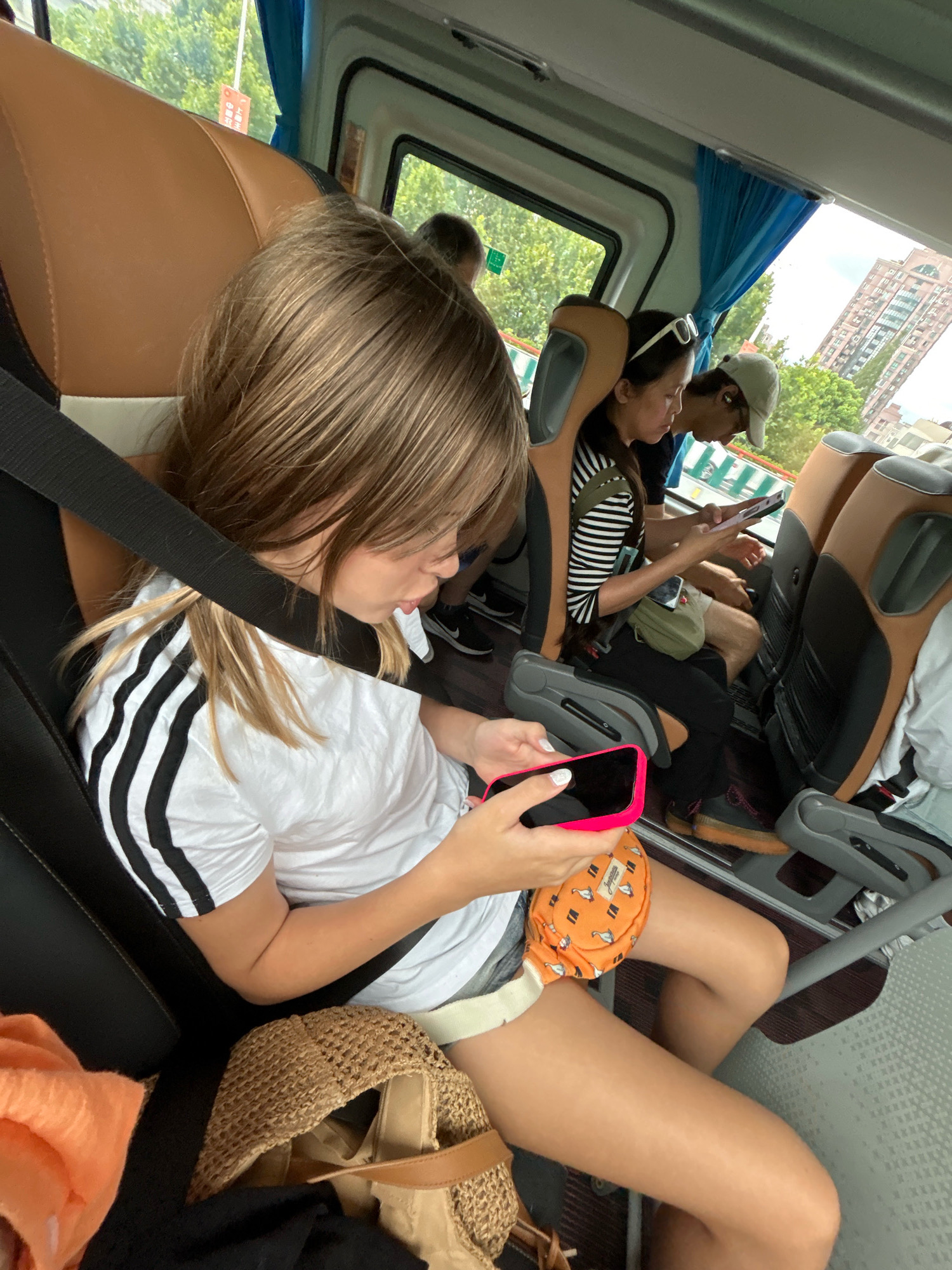 In the city of Changning District, China, the image captures the interior of a bus with several passengers. A young girl in the foreground, wearing a white t-shirt with black stripes on the sleeves, is seated and looking at her pink-cased smartphone. She is wearing a seatbelt, and her legs are visible, along with a straw bag and an orange pouch on her lap. Behind her, a woman with white sunglasses perched on her head is also looking at her phone, while a man wearing a cap and headphones is beside her, also engrossed in his device. The bus seats are upholstered in brown and gray fabric, and the windows offer a view of trees and buildings outside. The overall scene depicts passengers using their mobile devices during a bus journey.