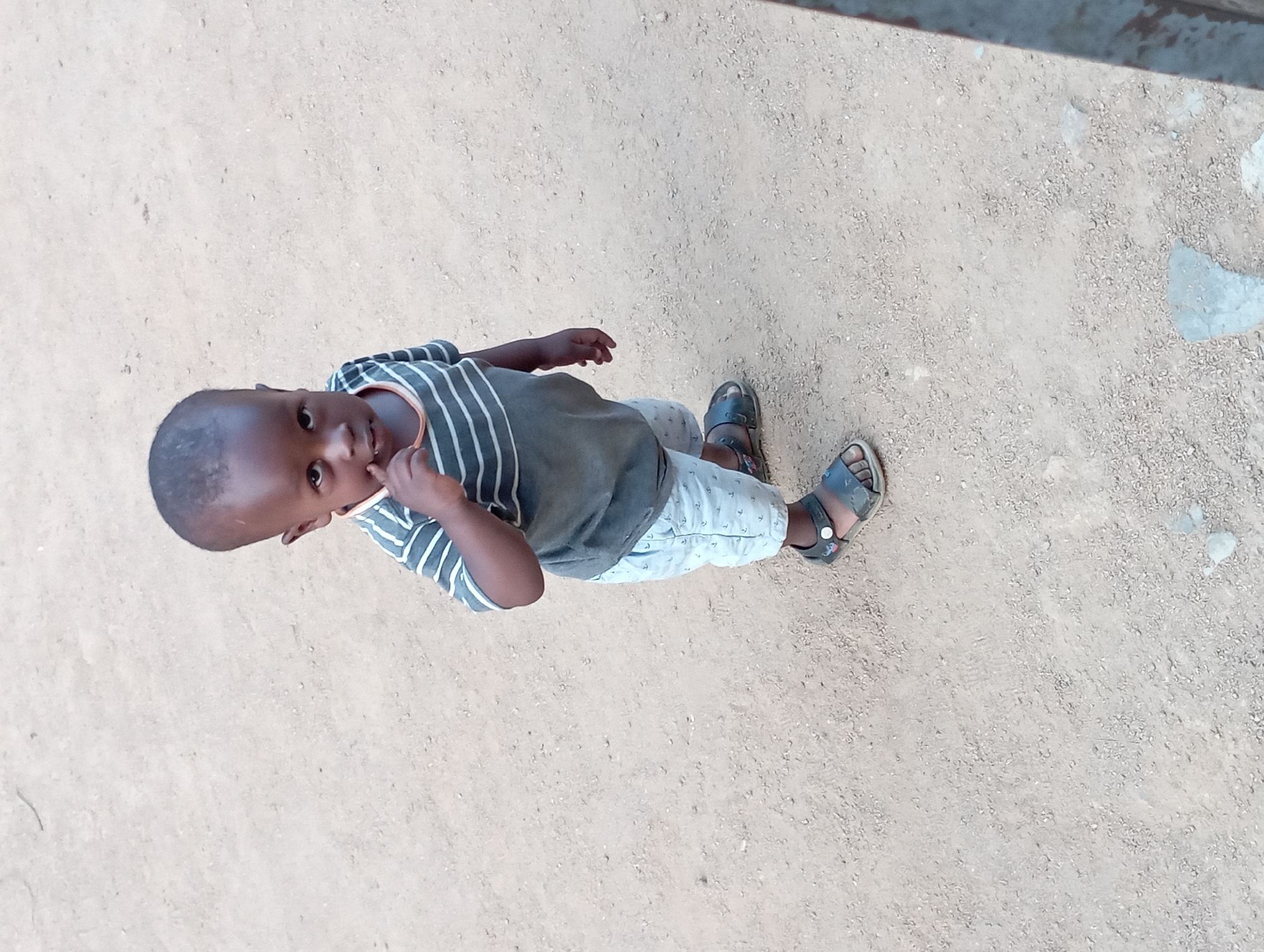 A young child is depicted standing outdoors on a light-colored, sandy or gritty ground. The photograph is taken from a high angle, looking down at the child who is gazing upwards towards the camera. The child wears a grey t-shirt with horizontal white stripes, light-colored shorts patterned with small anchors, and dark sandals. One of the child's fingers is positioned near their nose or mouth. The ground appears uneven and dusty, suggesting an unpaved outdoor area. The lighting indicates it is daytime. The child's expression is neutral, and they are directly engaging the camera. No text is visible in the image. The setting aligns with an outdoor environment in a location such as Jos, Nigeria.