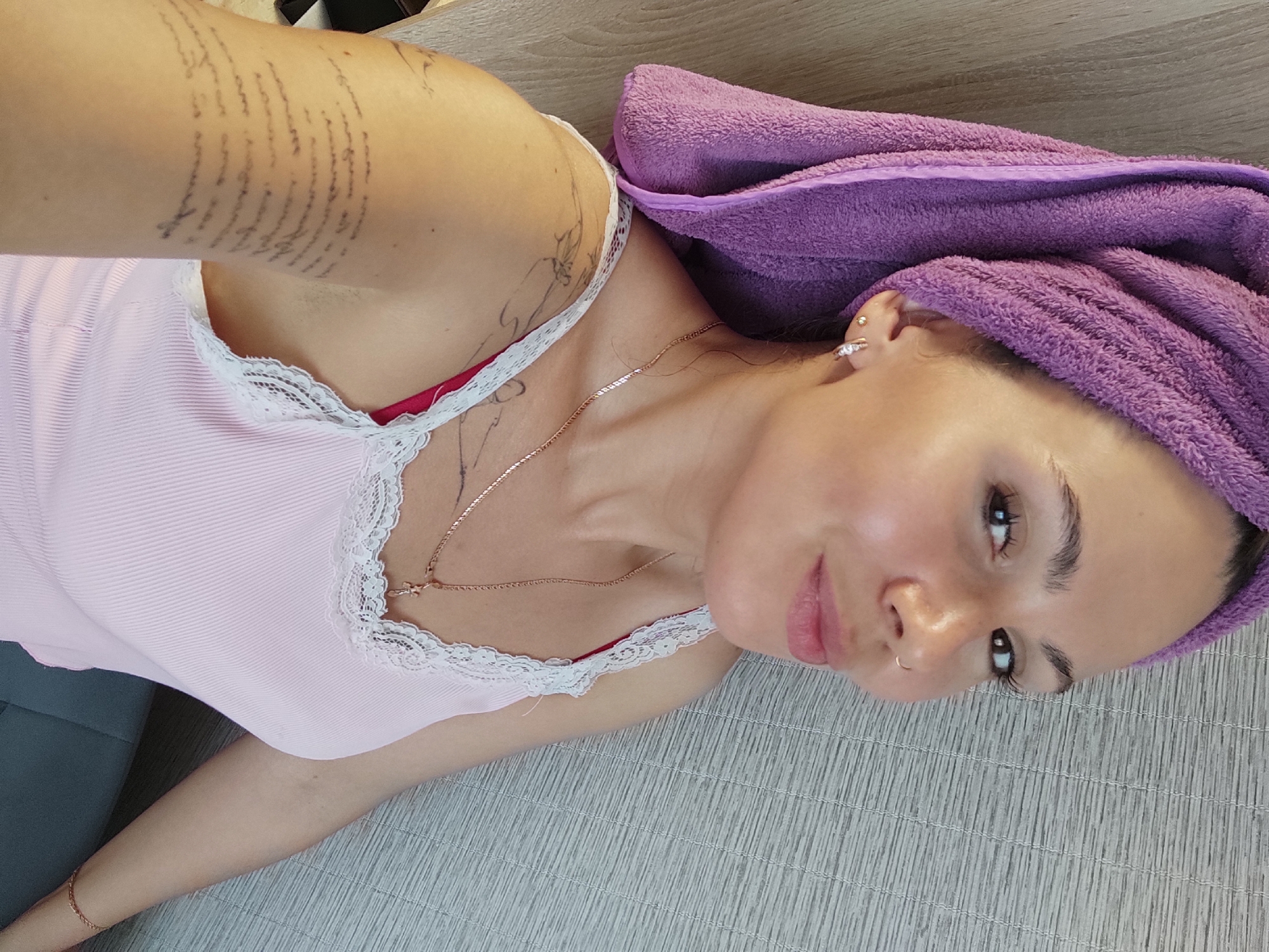A woman with a purple towel wrapped around her head is lying on a light-colored surface. She is wearing a light pink top with lace trim and a gold necklace. She has a nose ring and a tattoo on her arm. Based on an analysis of publicly available geotagged images, the photograph was taken in Chaikovsky, Russia.