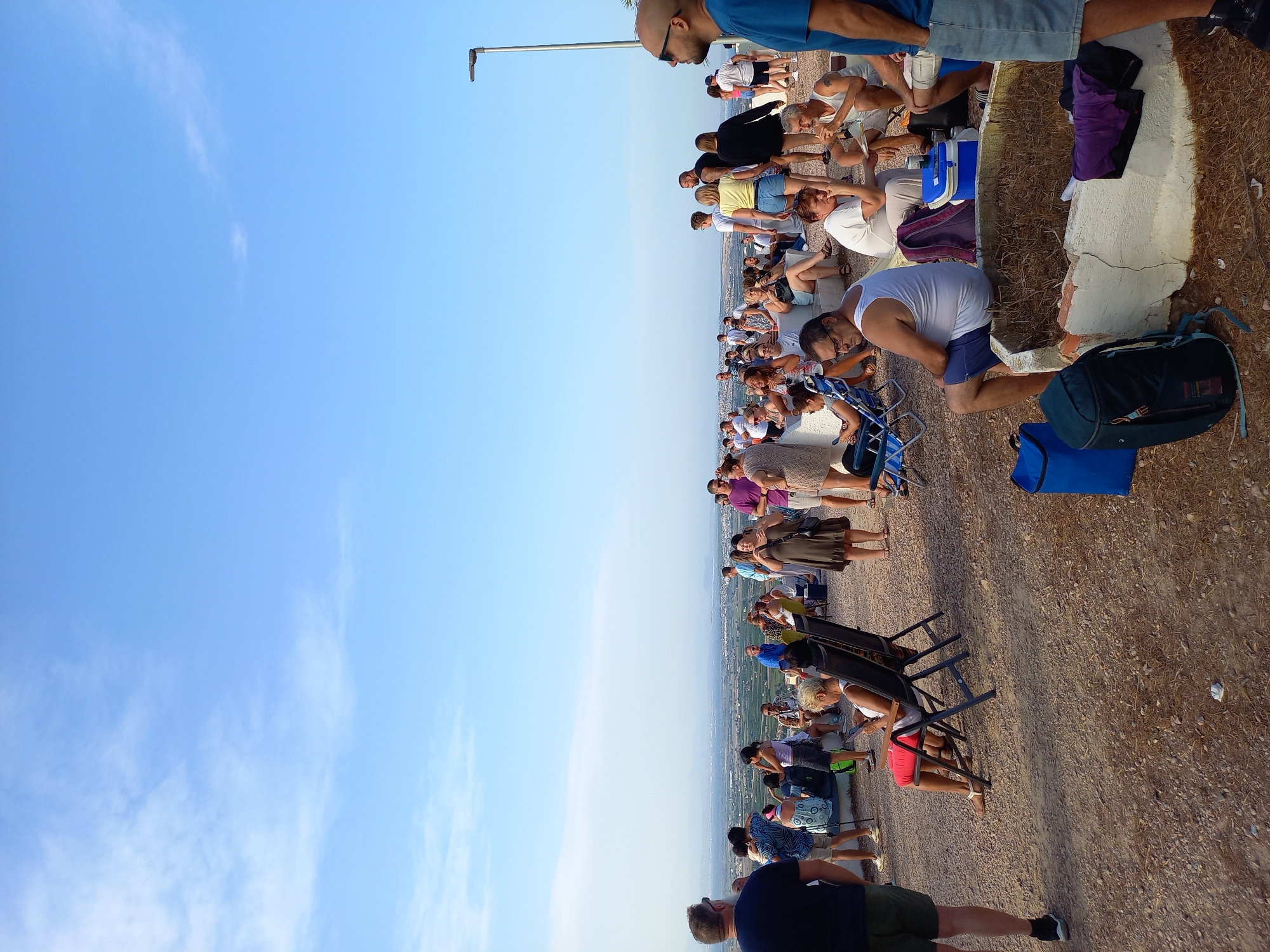 The image depicts an outdoor gathering of people on a hilltop overlooking a semi-arid landscape with buildings and vegetation. The sky is clear blue with sparse white clouds. A lamppost is visible on the right side of the frame. The ground is covered with gravel and dirt.
There are approximately 50-60 people in the scene, mostly adults, dressed in casual summer clothing. Many are standing or sitting in portable chairs. Some individuals are interacting, talking or looking at their phones. Objects include backpacks, a cooler, and a large stone planter filled with dried grass and debris.
The overall atmosphere appears relaxed, possibly a social event or a scenic viewpoint visit. The location is identified as Urbanización Ciudad Quesada, Spain.