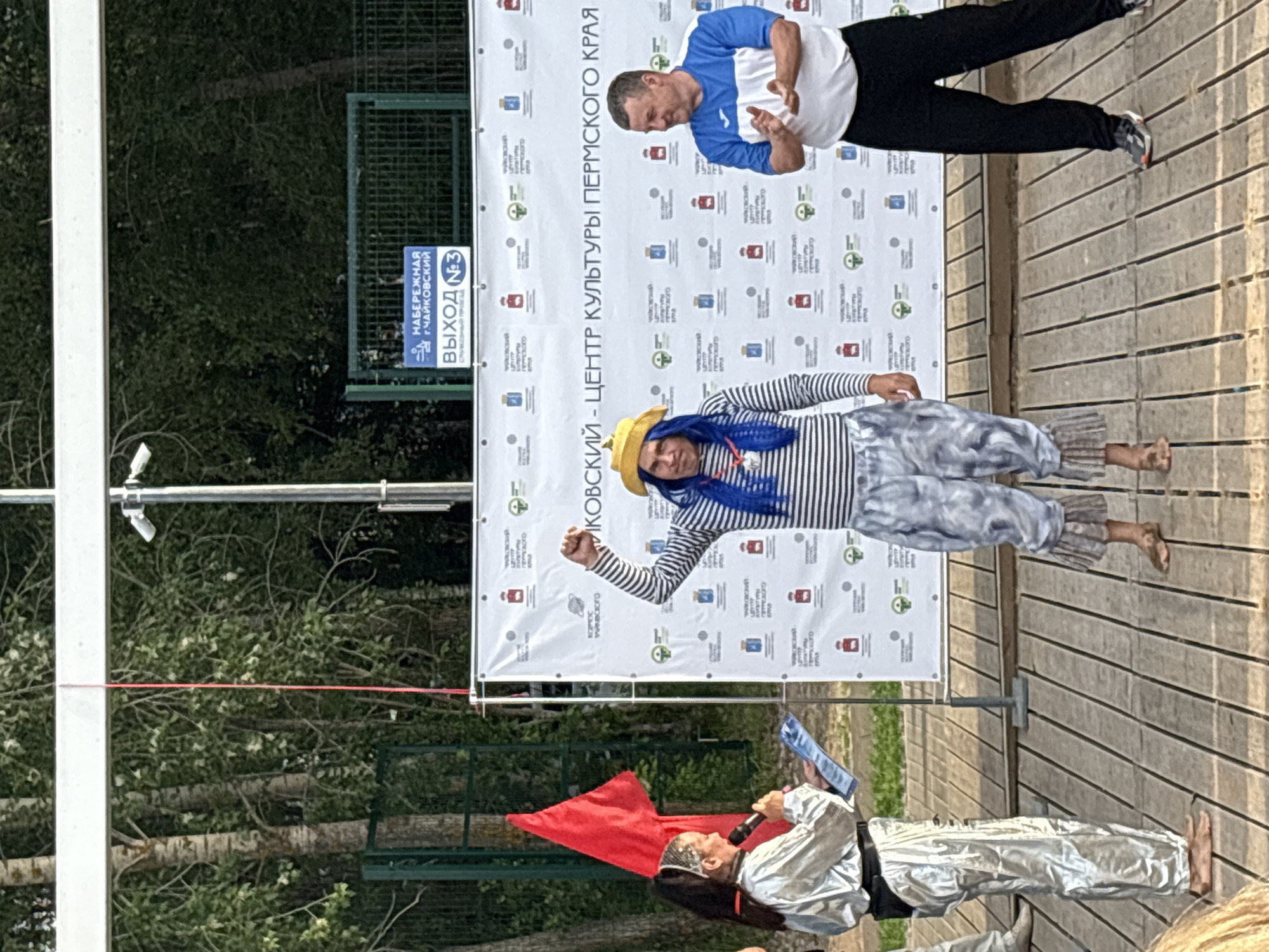 The image shows an outdoor event on a wooden stage in Chaikovsky, Russia, likely near the Naberezhnaya (Embankment). Three people are visible. On the left, a woman in a silver, shiny outfit with a black belt and black headband is speaking into a microphone and holding a document. To her right, a person in a pirate-like costume with a yellow hat, blue wig, striped shirt, and baggy pants is raising their right fist. On the right, a man in a white and blue athletic shirt and black pants is clapping. Behind them is a large banner with Russian text and logos, and a sign indicating "NABEREZHNAYA G. CHAYKOVSKY" (Embankment, Chaikovsky City) and "VYKHOD №3" (Exit No. 3). Trees and a green fence are visible in the background.