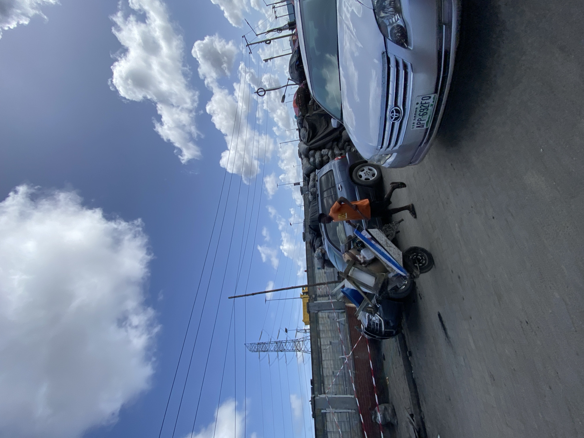 The image depicts a street scene in Lagos, Nigeria. The sky is blue with scattered white clouds. Overhead power lines are visible. The street is lined with buildings, some with red and white striped barriers.
A silver Toyota car is parked on the right side of the frame, with a license plate that reads "APP-692FO". Adjacent to the Toyota, a man wearing an orange shirt appears to be working near or underneath a vehicle, possibly a blue truck or van. The bed of this vehicle appears to be overloaded with materials, possibly sacks or other goods.
There are other cars and objects along the street, suggesting a bustling urban environment. The overall impression is that of a busy commercial or residential area in Lagos.