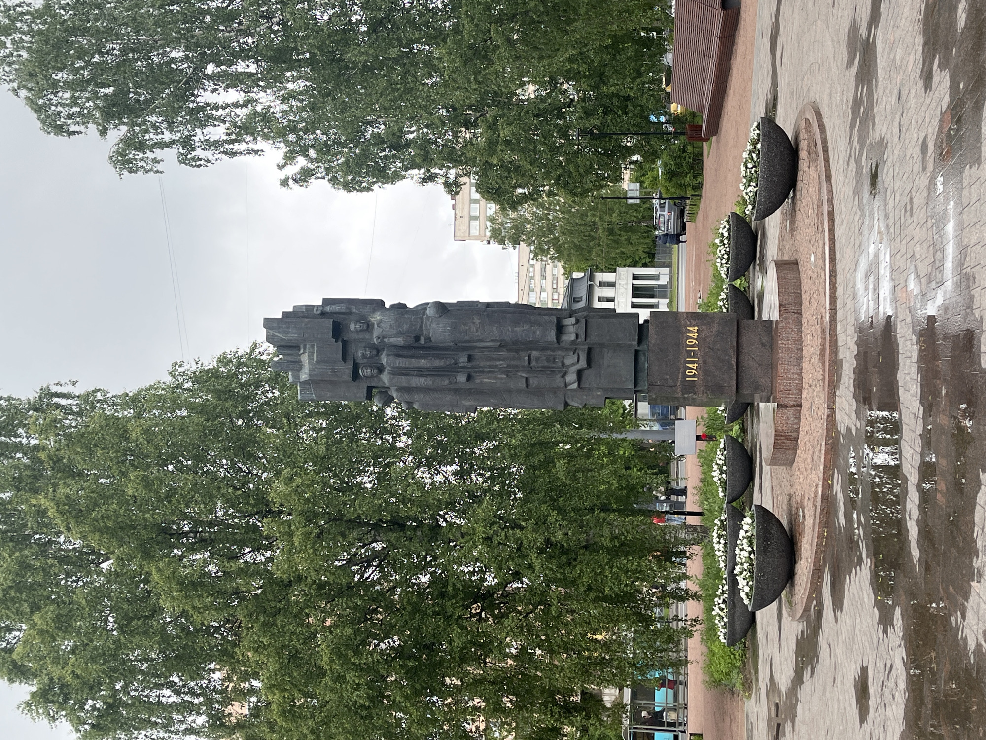 The image shows the Motherland Monument located on Piskaryovskoye Memorial Cemetery in the city of Saint Petersburg, Russia. This cemetery serves as the burial place for approximately 500,000 victims of the Siege of Leningrad during World War II, from 1941 to 1944, as inscribed on the monument. The monument is a dark stone sculpture depicting a group of figures. The ground around the monument is paved with stone, and several half-spherical flower pots filled with white flowers are positioned around the base. Trees frame the monument, suggesting it is located in a park-like setting. The weather appears overcast, and the ground is wet. In the background, buildings are visible, indicating the memorial is located within an urban area.