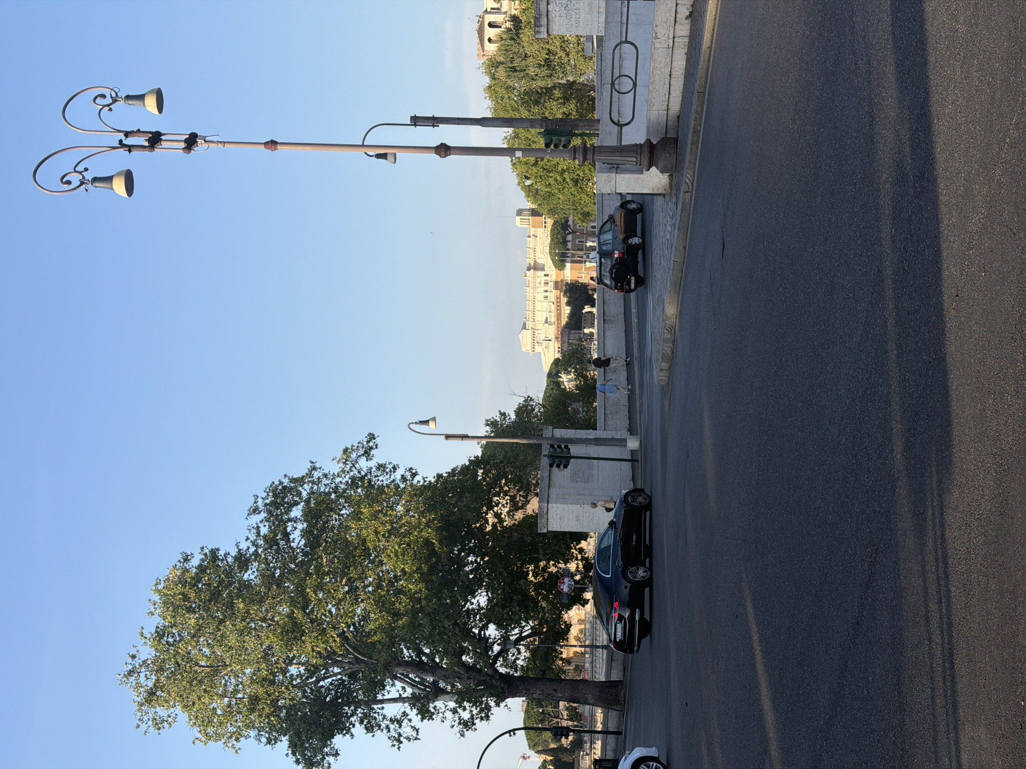 This is a wide shot of a street in an urban environment, possibly Rome given the architectural style and foliage. The sky is clear and blue. A large, leafy tree is on the left side of the frame, casting shadows on the asphalt road. In the middle ground, there is a white stone bridge with ornate streetlights. Several cars are visible on the road, including a black sedan and a black hatchback. Two people are walking on the sidewalk. In the background, there are buildings with light-colored facades and trees. The overall impression is a bright, sunny day in a city.