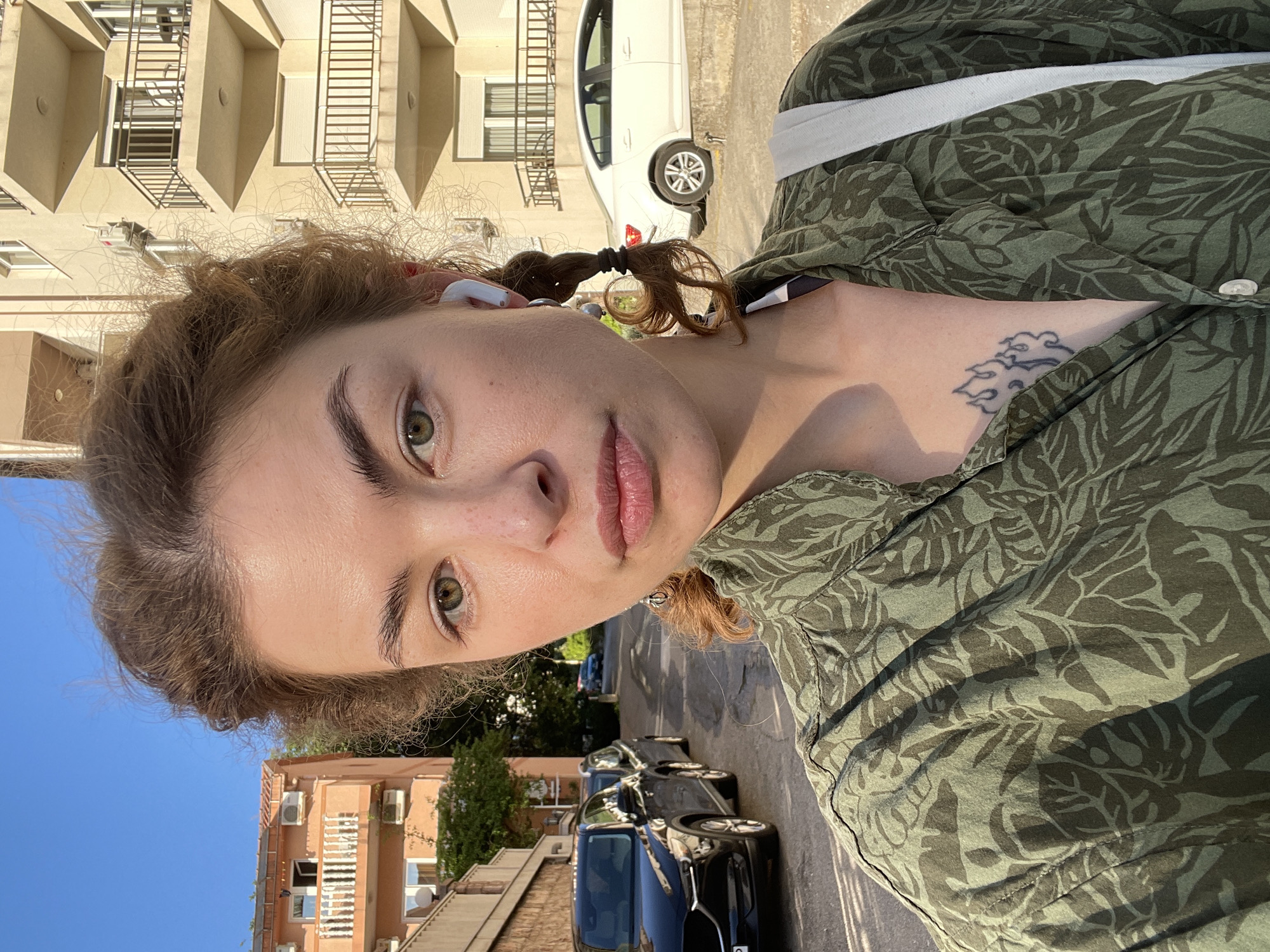 A young woman with green eyes and auburn hair is seen in a selfie-style image. Her hair is styled in two pigtails, and she is wearing a white earbud in her left ear. She is wearing a button-down shirt with a green leafy pattern. A tattoo is visible on her upper chest/shoulder area. Behind her, there is a street scene with cars parked on the side of the road. Buildings are visible on both sides of the street. The specific location is Rafailovići, Montenegro. The time of day appears to be during daylight hours, based on the lighting.