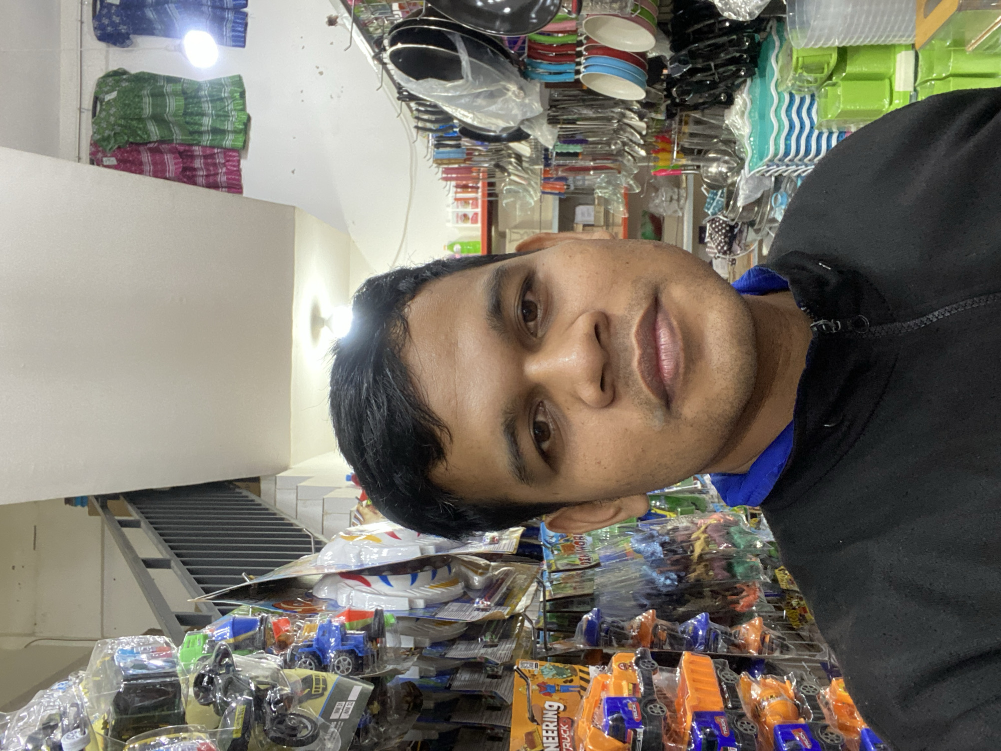 The media file is a selfie taken in what appears to be a retail store, possibly in Lamteumen Barat, Indonesia. The subject is a man with dark hair and brown eyes, facing the camera. He is wearing a black hooded jacket with a blue undershirt. His expression is neutral.
The background is filled with shelves of various products. On the left side of the image, stacks of what appear to be folded textiles in blue, green, and pink patterns are visible. Above these, a white wall is present, and to the right, a metal staircase is visible.
Further to the right, the shelves are packed with a variety of items. Some are clear plastic containers, others are colored kitchenware like plates and bowls in red and blue. There are also many toys, predominantly toy trucks and construction vehicles, some packaged in plastic. The overall impression is of a crowded retail environment selling household goods and toys.
The lighting in the scene seems to be artificial, with bright light sources illuminating the products. There is no indication of the time of day or weather conditions. The presence of a wide array of consumer goods suggests the setting is a shop or market.
Visible text includes "ENGINEERING TRUCK" on the packaging of some of the toys. The orientation of the image is rotated.