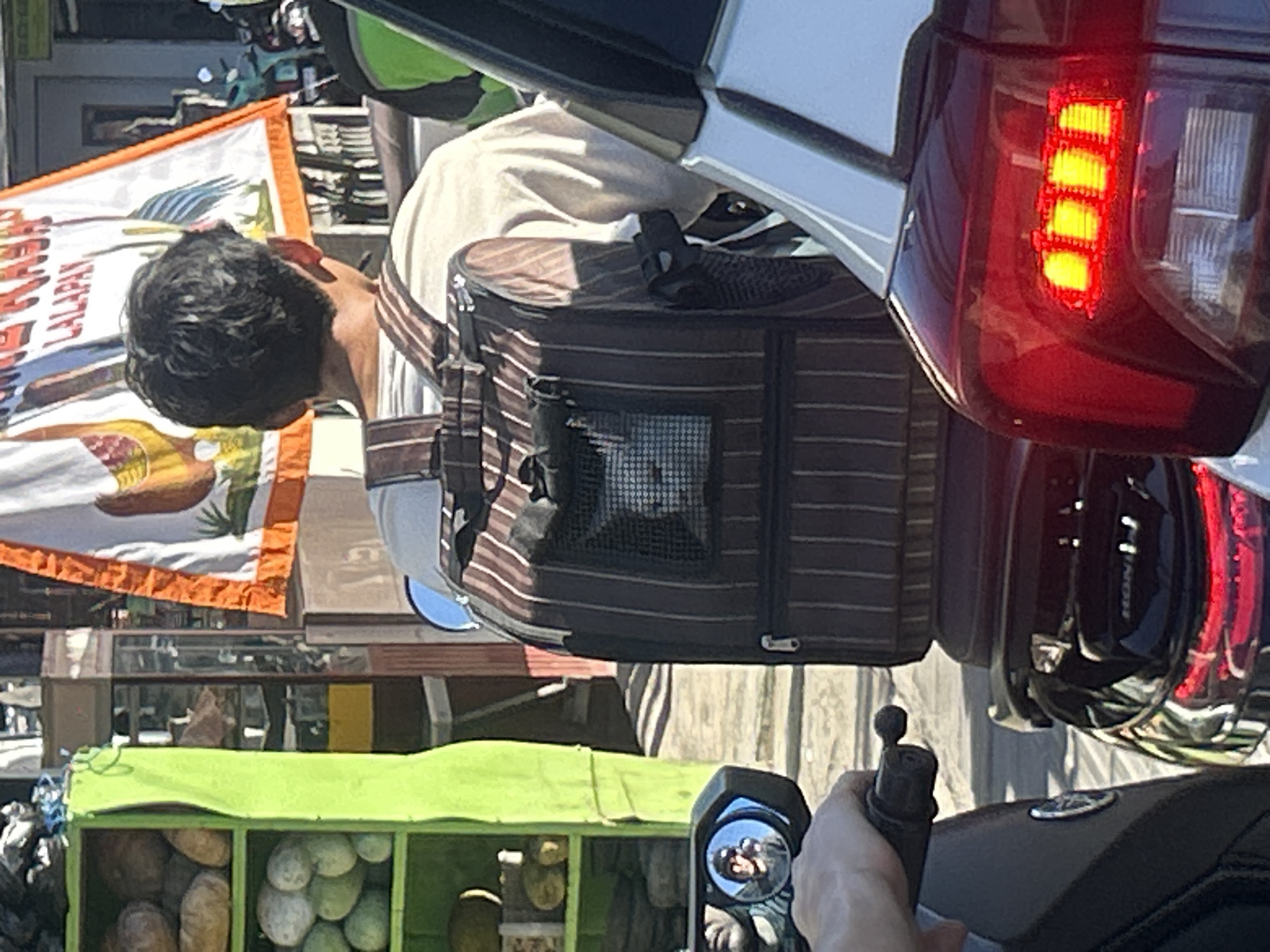 The image, taken from the perspective of a person riding a motorcycle, shows a man on another motorcycle transporting a striped pet carrier behind a white car. The car's red taillight is prominent in the frame. The scene takes place in North Kuta, Indonesia, as indicated by the local flag with the inscription 'LALAPAN CABANG', which is hanging from a food stall. The green food stall displays various melons. The motorcycle mirror reflects the photographer.
