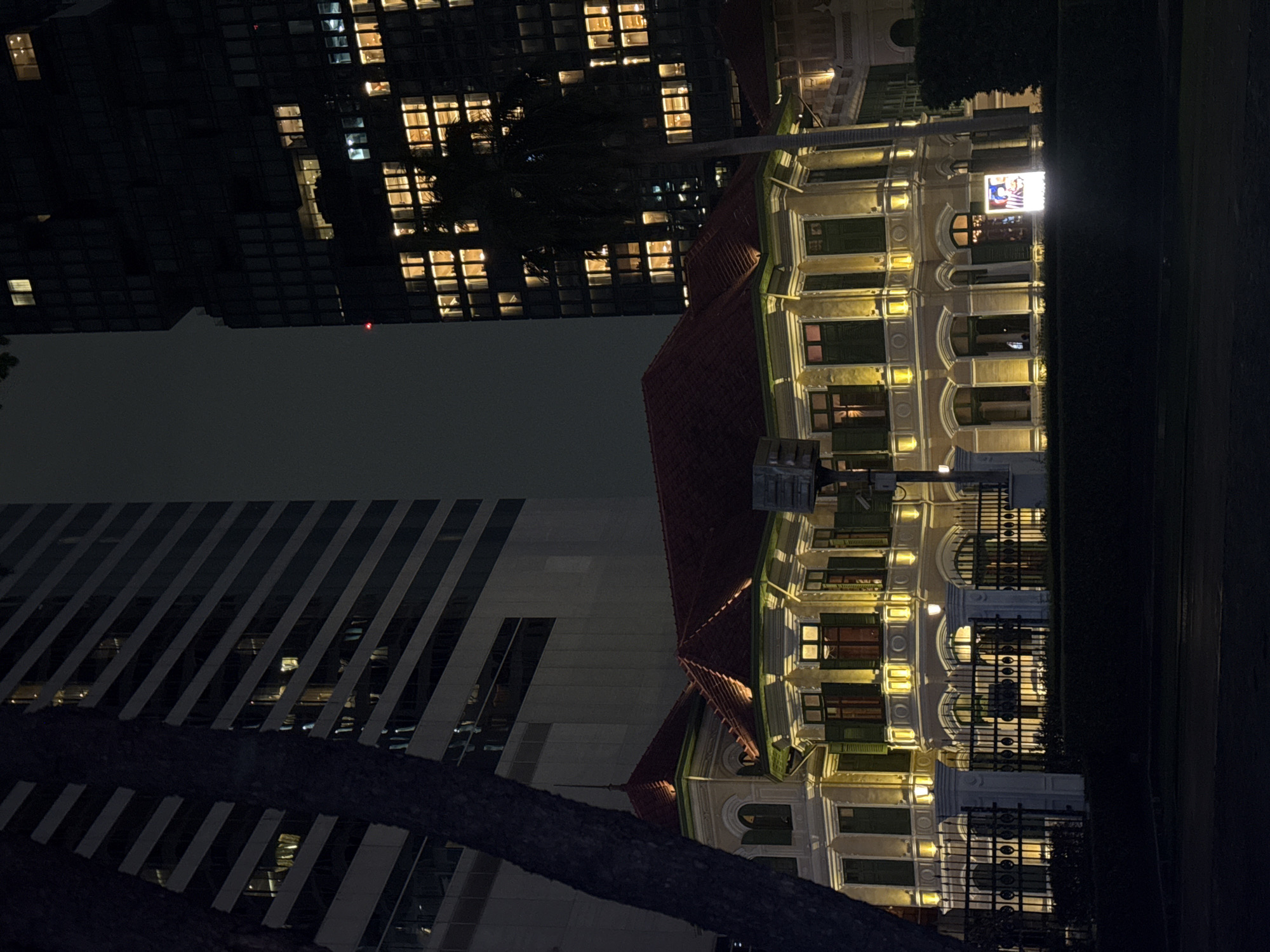 The image depicts a historical building illuminated at night in Bangkok, Thailand. The building is a light-colored, two-story structure with a red tiled roof, arched windows with green shutters, and ornate architectural details. It is surrounded by a black metal fence and hedges. Behind the historical building, two modern skyscrapers are visible. The left skyscraper is tall and has horizontal lines, with some windows lit. The right skyscraper is darker and appears more angular, with numerous illuminated windows. A palm tree stands in front of the right skyscraper, partially obscuring the historical building. The scene is set at night, with the lighting focusing on the historical building, creating a contrast between the old and the new architecture. The sky is dark, suggesting it is nighttime. There are no people clearly visible in the image.