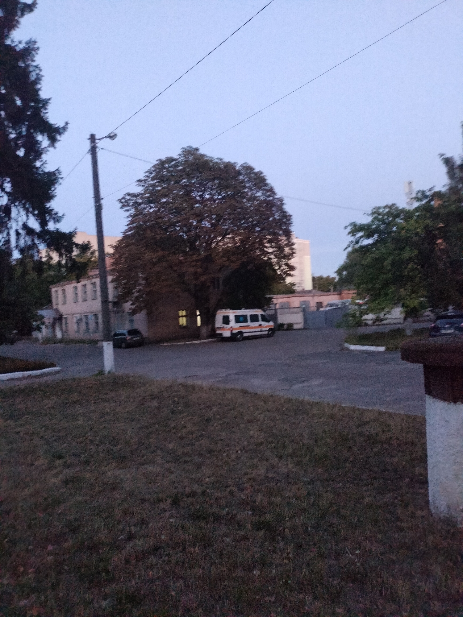 A slightly blurry, low-light exterior scene in Chernihiv, Ukraine, features a building with several windows, a large tree with dark foliage, and an ambulance parked in the lot. The building appears to be of moderate size, possibly a hospital or administrative facility, with a light-colored facade and some visible architectural details. The ambulance is white with red and blue markings, indicative of an emergency medical vehicle. A dark-colored car is parked near the building. In the foreground, a grassy area with dry grass is visible, suggesting it may be late summer or early autumn. Overhead, power lines are strung between poles. The sky is a pale, clear blue, indicating either dawn or dusk. The overall impression is one of a quiet, institutional setting.