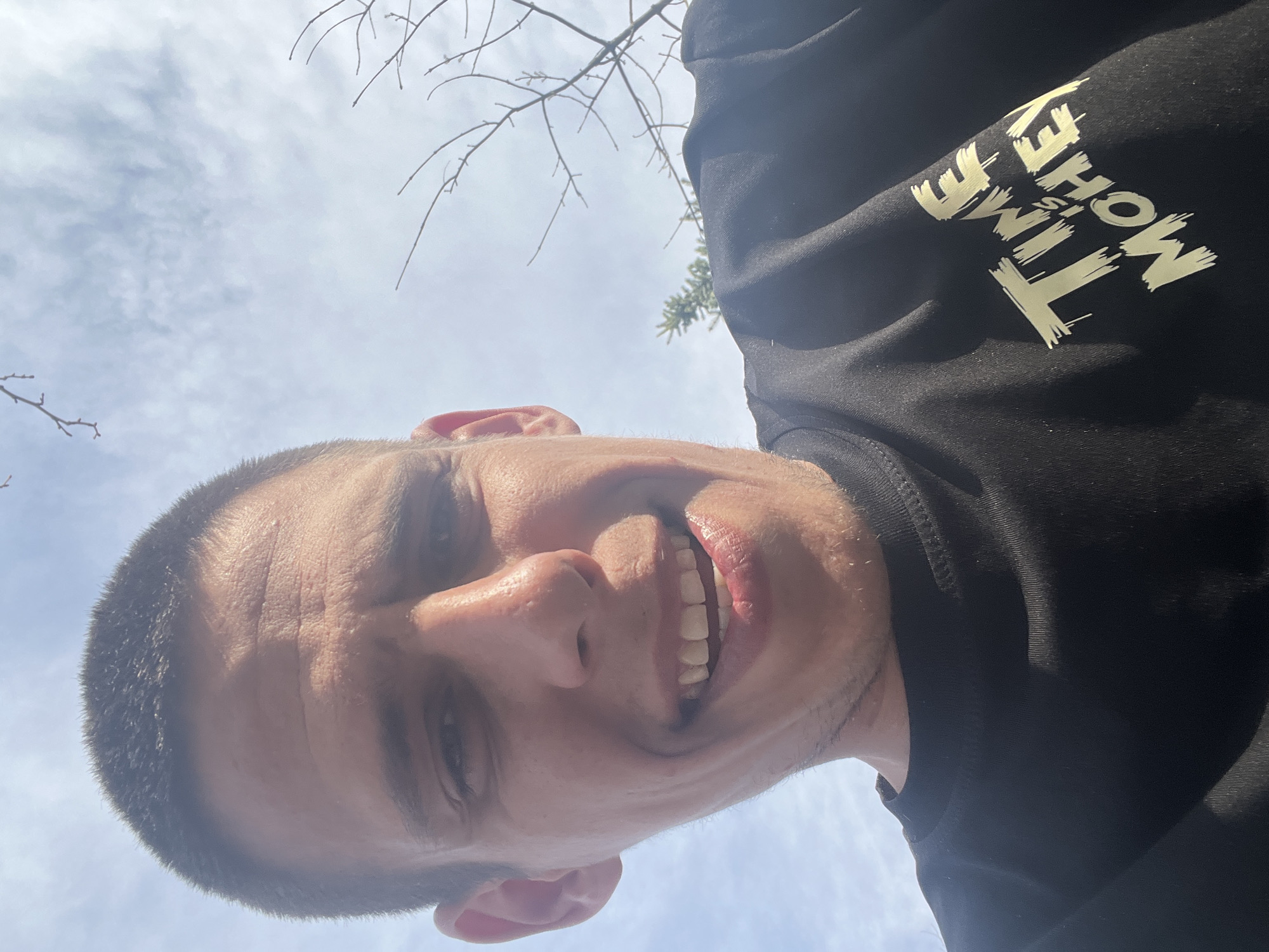 A man takes a selfie outdoors, looking up at the camera. He is wearing a black sweatshirt with the words "TIME IS MONEY" printed in yellow. The sky is visible in the background, with some clouds and tree branches. The location is indeterminate; there is no way to know if this photo was taken in Sofia, Bulgaria.