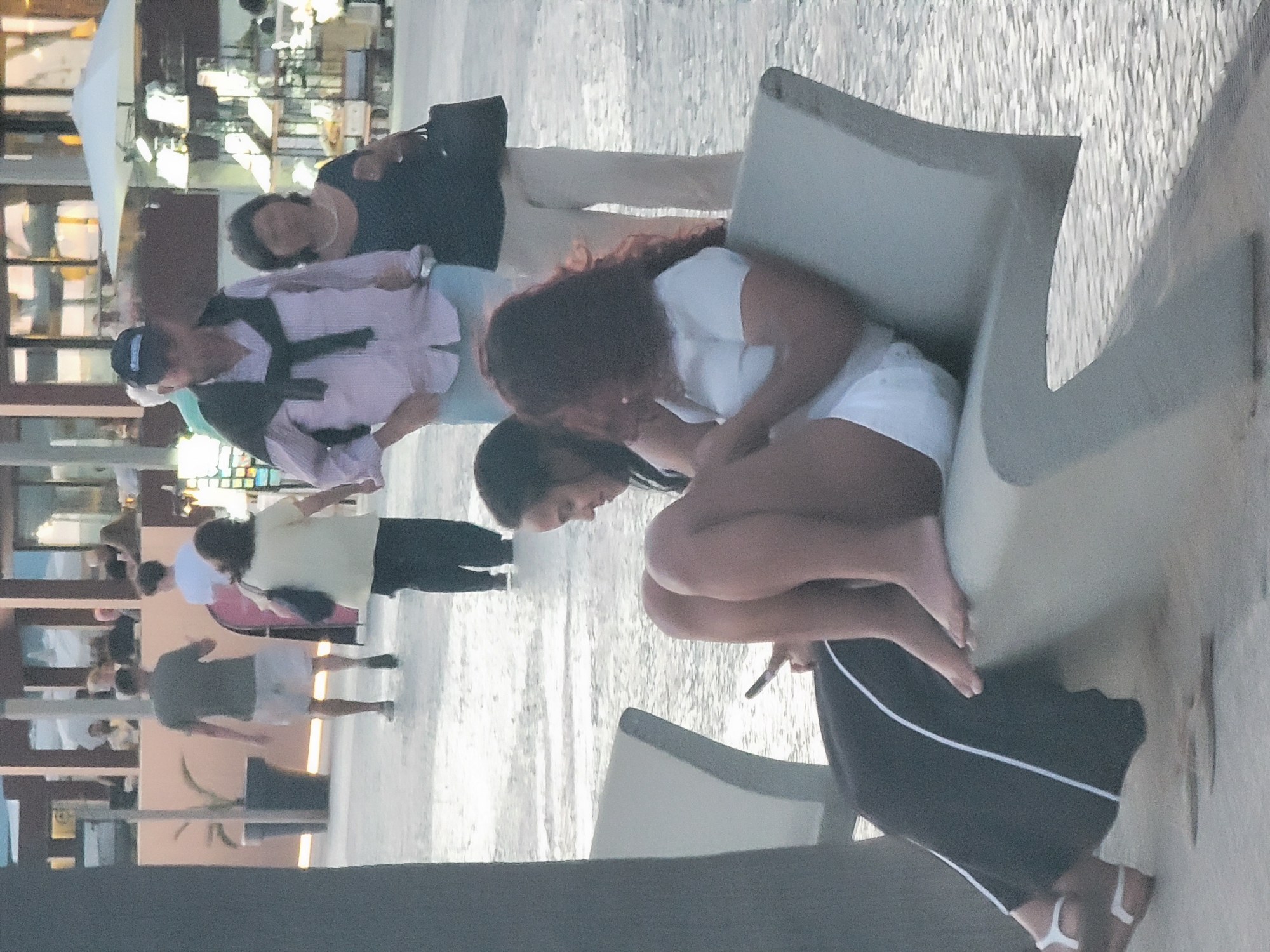 The image depicts a daytime scene along a promenade in Quarteira, Portugal. In the foreground, two women are seated on a modern, light-colored bench. The woman on the left is mostly obscured by the woman on the right, but is wearing dark, loose-fitting trousers with a white stripe down the side and white sandals. The woman on the right is wearing white shorts and a white t-shirt, with her legs crossed. She is holding a smartphone in her right hand, and her left hand is resting on her lap.
In the middle ground, a man in a light pink, button-down shirt and blue shorts is walking towards the right, with a dark sweater draped over his shoulders. He is wearing a baseball cap. To his left and slightly behind him, a woman in a light-colored top and dark trousers is walking in the same direction. She is carrying a black handbag. Further to the left, a man in a dark t-shirt and light-colored shorts is walking away from the camera. He is partially obscured by a large potted plant and a vertical pole.
The background shows building facades with large windows, suggesting shops or restaurants. There are also market stalls visible, with an umbrella providing shade. The ground is paved with light-colored cobblestones. The overall atmosphere appears casual and commercial, consistent with a public walkway or shopping area in a coastal town.