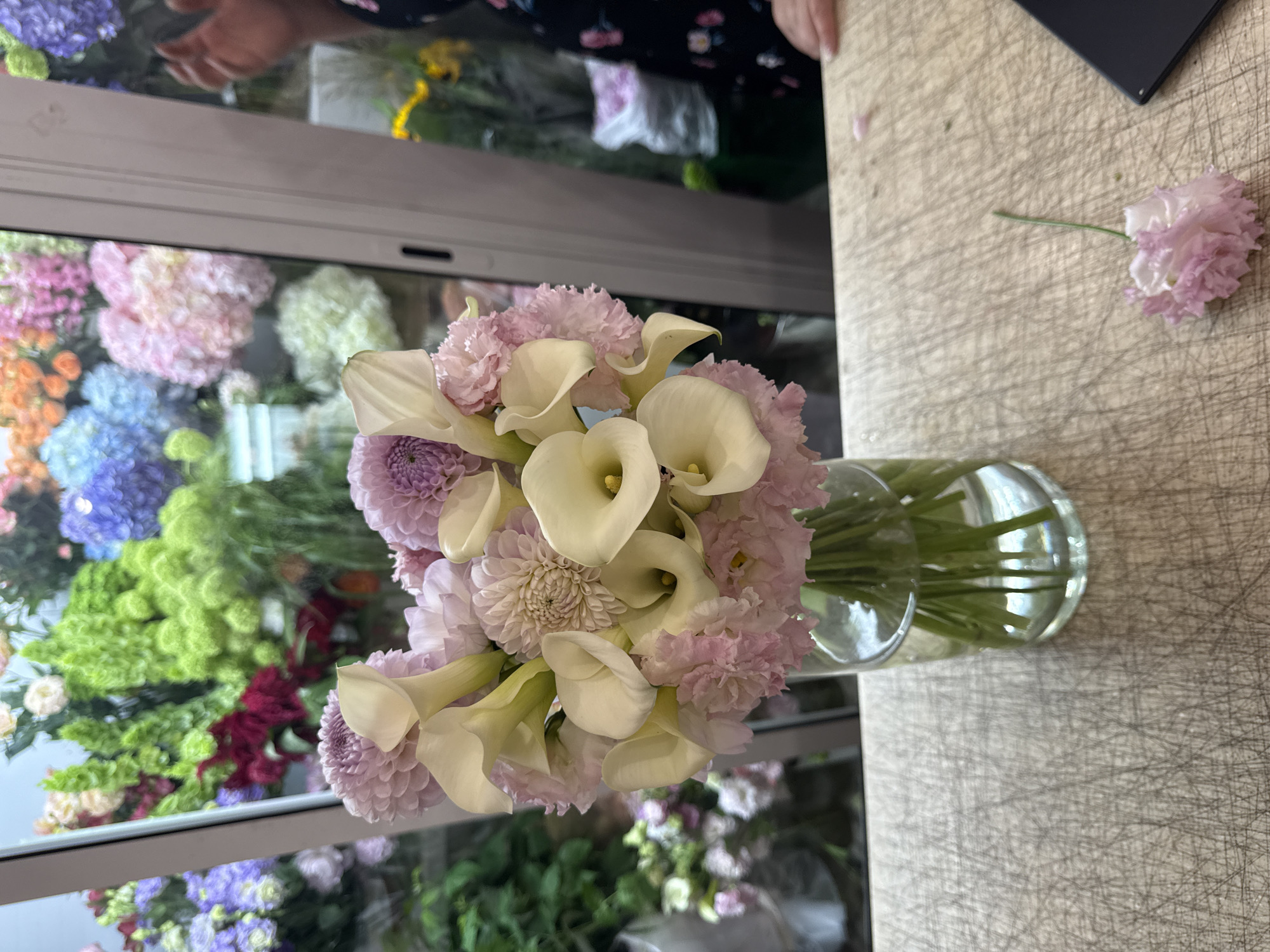 A close-up shot focuses on a bouquet of white calla lilies and pink carnations in a glass vase filled with water. The flowers are arranged on a wooden surface that appears to be a cutting board. The background is blurred but shows a colorful display of various flowers, suggesting a floral shop. A person's hand is visible on the right side of the image, reaching towards the flowers. The scene appears to be set indoors, with natural light illuminating the flowers from what seems to be a window. The overall impression is one of fresh flowers and a professional setting. The image does not contain any information that would allow it to be located in Assinovskaya, Russia.