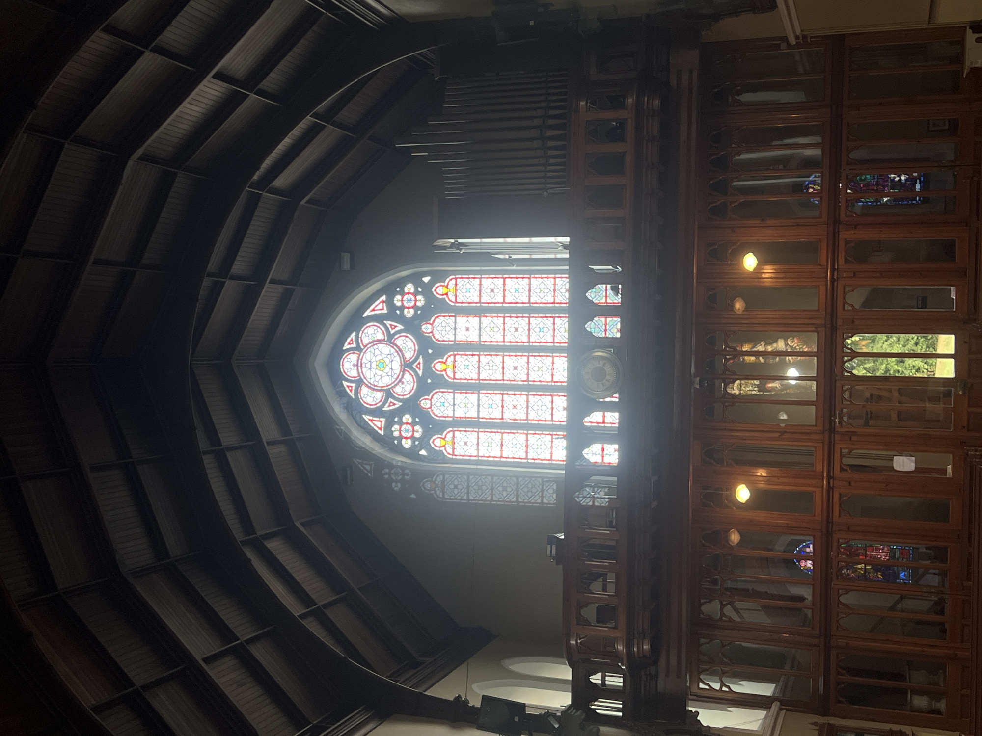 A medium-shot photograph captures the interior of a dimly lit church, showcasing its architectural features and stained-glass window. The vaulted wooden ceiling with exposed beams dominates the upper portion of the image. Below the ceiling, a large, ornate stained-glass window is prominently displayed, featuring intricate geometric patterns and vibrant red and yellow colors, with a circular motif at the top.
In the mid-ground, a wooden balcony or choir loft with decorative railings spans across the width of the church. Beneath the loft, a multi-paneled wooden structure with gothic arch detailing is visible. Various stained-glass elements can be seen through the panels, along with what appears to be an organ on the right side of the frame. The lower part of the image reveals more wooden paneling and seating areas, with glimpses of greenery through a window on the far left. The overall impression is one of a grand, historic ecclesiastical space.
