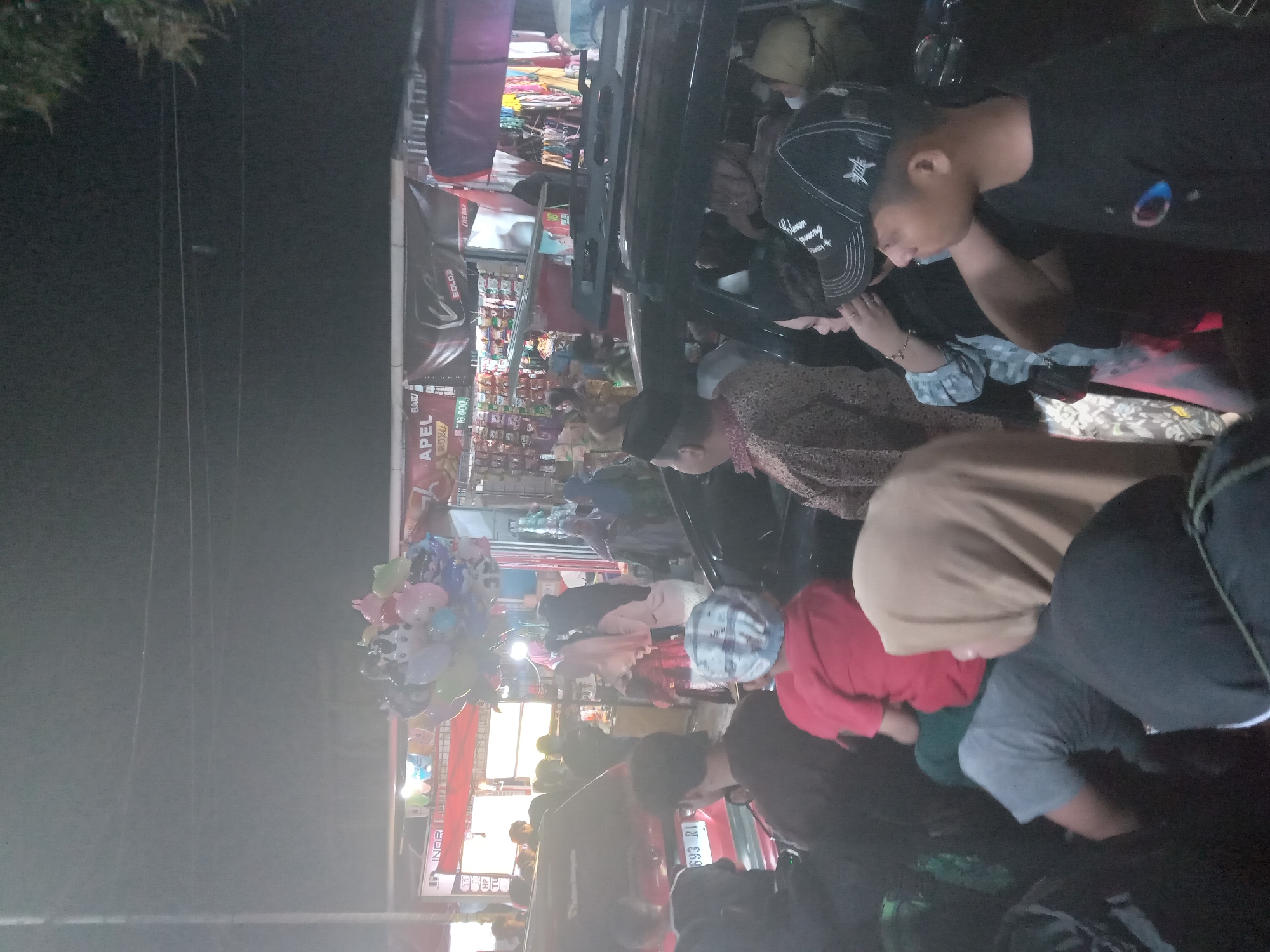 An overhead shot depicts a dimly lit outdoor gathering at night, likely a street market or festival. In the foreground, several people are visible, with a focus on those in vehicles. A black SUV with a roof rack is partially obscured by the people standing around it. A red car with a license plate reading "693 RI" is also present. The people in the foreground are diverse in age and attire, with some wearing hats and headscarves. Some appear to be interacting with each other, while others are looking in different directions. In the background, stalls are set up with various items for sale, including balloons. The overall impression is one of a lively, crowded public event.