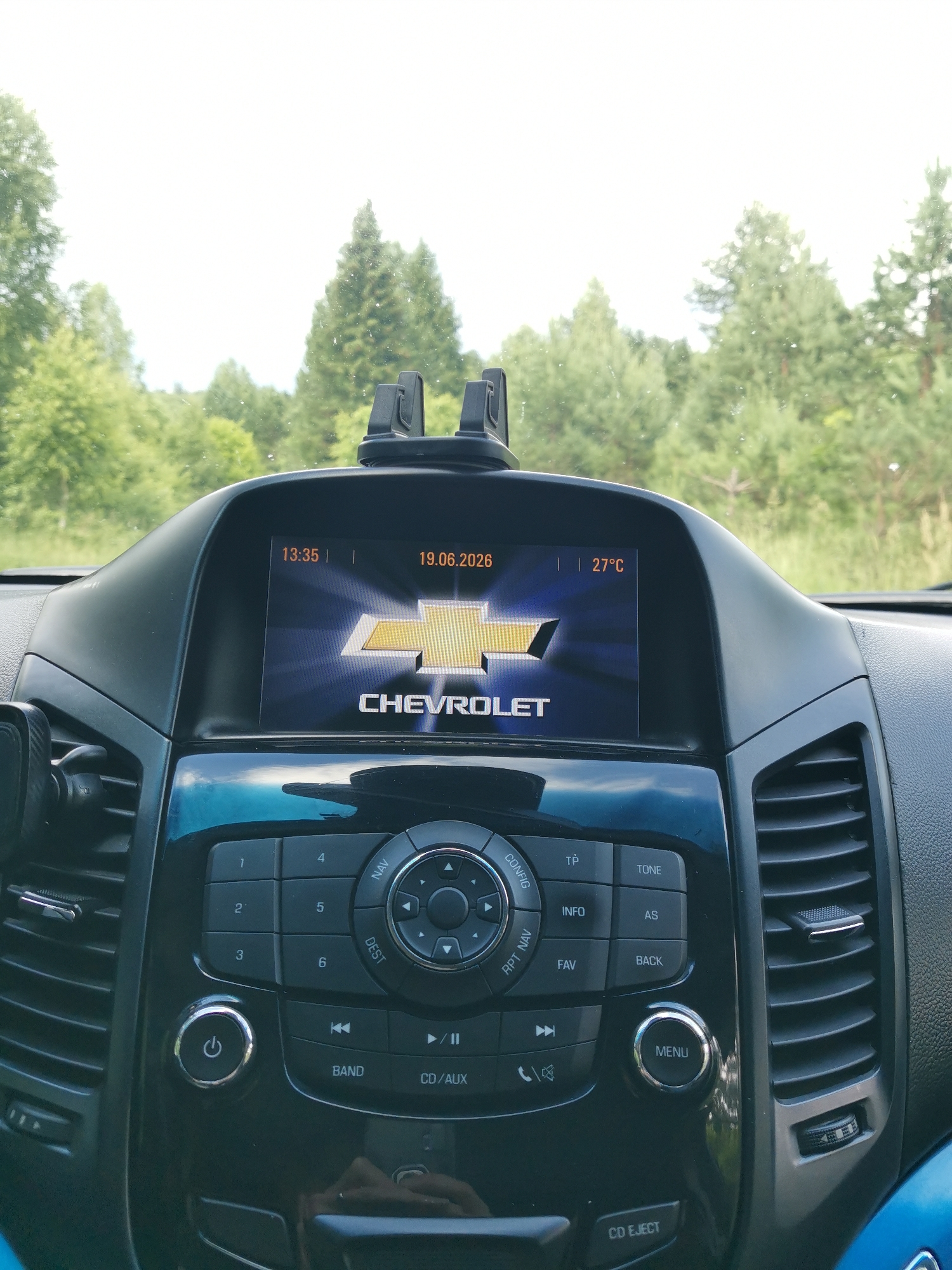 The image shows the interior of a Chevrolet car, with a focus on the dashboard infotainment system. The screen displays the Chevrolet logo, the time "13:35," the date "19.06.2026," and the temperature "27°C." Below the screen are various buttons and controls for the audio system, including numbered presets, navigation, audio settings, and a CD/AUX input. A power button is visible to the left, and a menu button is to the right. The car appears to be parked or driving outdoors, with a blurred background of green trees and a bright sky visible through the windshield.