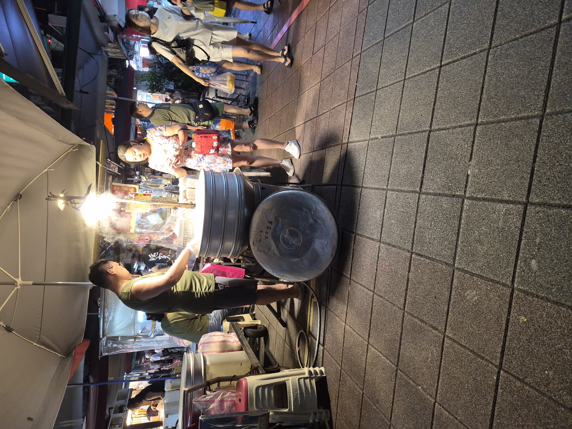 The image depicts a street scene in Taipei, Taiwan, likely a night market given the stalls and lighting. The foreground shows a paved ground of square tiles. In the midground, a man in a green tank top is tending to a large steamer, with steam rising from it. He is wearing shorts and glasses. Next to him, a woman in a floral dress is looking at her phone, holding a small red bag. Behind them, a man in a dark shirt and shorts is visible. Further to the right, a woman in a white t-shirt and shorts is walking away, carrying a bag. The background shows more market stalls with various items and people. Overhead, umbrellas and lighting fixtures are present, creating a bustling night market atmosphere.