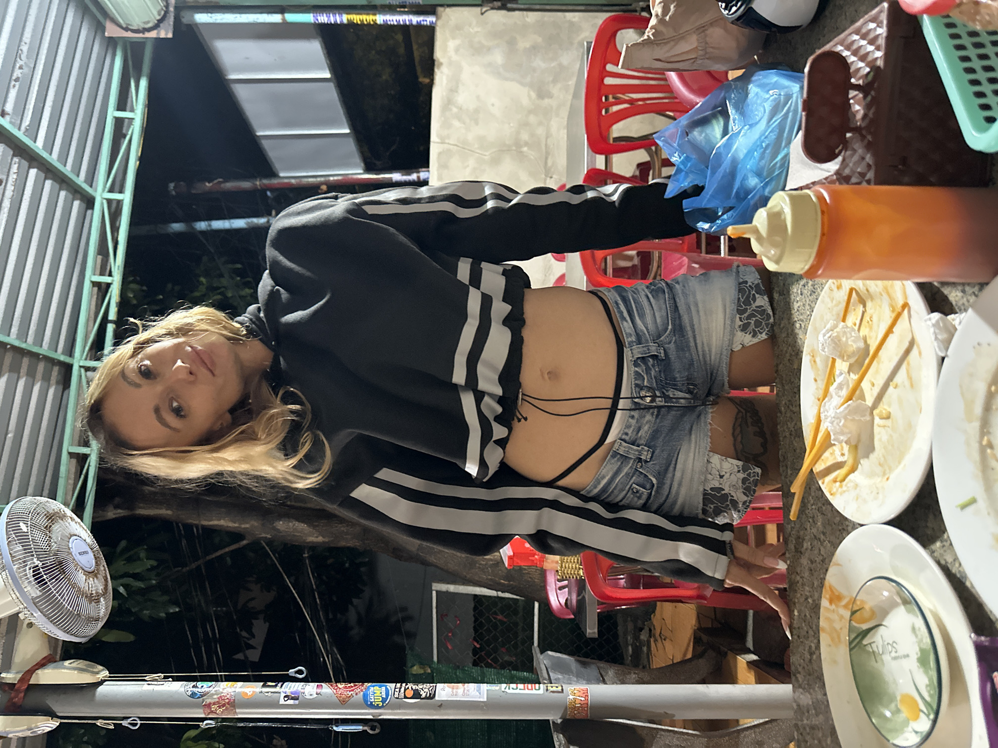 The image shows a woman at an informal outdoor or semi-outdoor eating area during the night. The woman, with long blonde hair, is looking towards the camera with a neutral expression. She is wearing a black cropped hoodie with white stripes on the sleeves, and denim shorts, revealing her midriff and a belly button piercing. A tattoo is visible on her upper right thigh.
She is positioned near a table cluttered with the aftermath of a meal. On the table, there is an orange squeeze bottle, likely containing a sauce, and several dirty white plates holding food remnants such as skewers and possibly fried items, along with a pair of chopsticks. A blue plastic bag and a brown woven container are also present on the table. Red plastic chairs are visible around the table, characteristic of casual dining establishments in Vietnam.
To the left of the frame, a white oscillating fan suggests warm weather. The background is dark, indicating night time, with some trees and foliage visible. The setting includes simple architectural elements like corrugated metal panels, concrete walls, and a metal support structure. A metal pole in the foreground is covered with various stickers, some featuring legible text like "JUMP" and "LODECK." One of the plates has the word "Tulips" printed on it, accompanied by a floral design. The scene evokes a casual street food or local eatery atmosphere, consistent with the provided location context of Phu Thuy Ward, Vietnam.