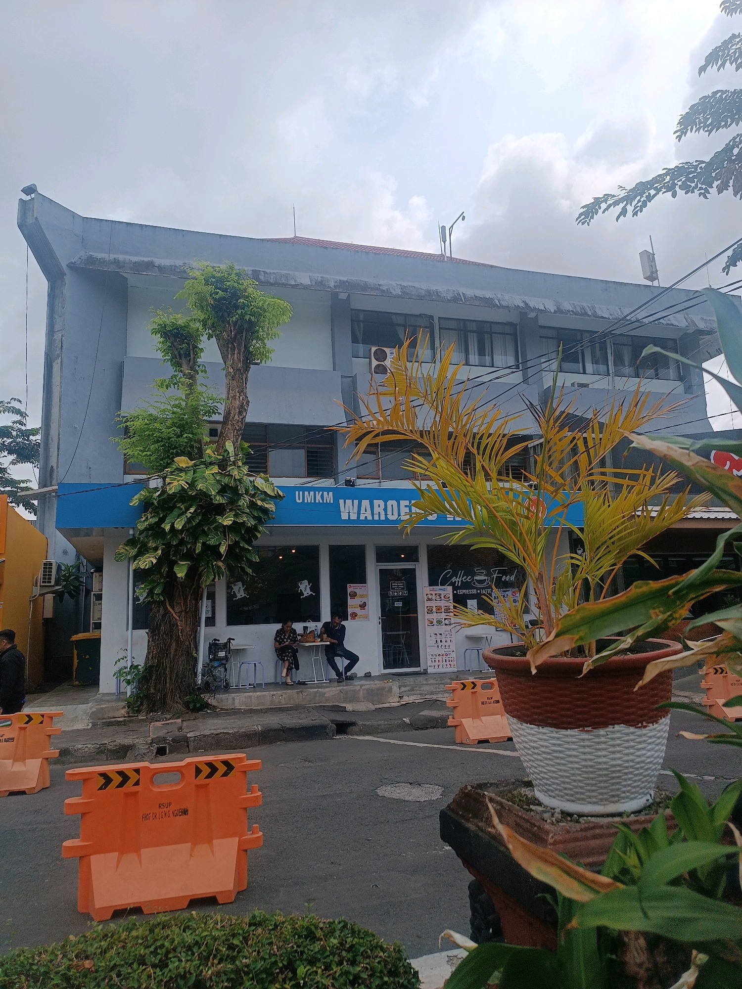 The image shows a building with a blue awning that says "UMKM WAROES" in white lettering. Below the awning, there are large windows that reveal a cafe interior with people sitting at tables. The building is light blue with dark gray accents and has multiple windows on the upper floors. In front of the building, there is a sidewalk with some trees and plants. Orange traffic barriers are placed on the road in front of the sidewalk. A potted plant with yellowing leaves is prominently displayed in the foreground. The sky is overcast, suggesting a cloudy day. The overall scene appears to be a street view in a commercial area.
