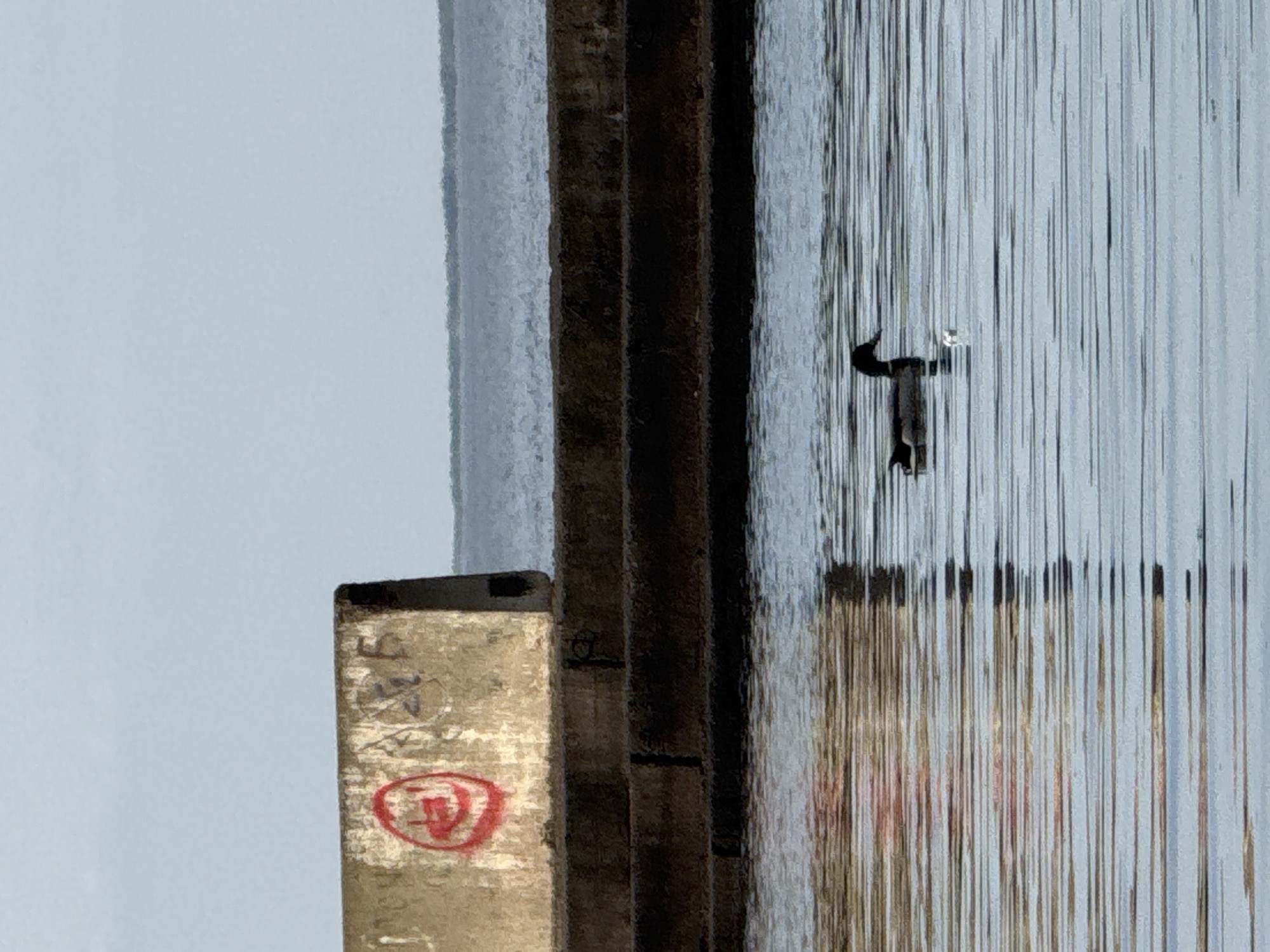 The image presents a close-up view of a section of an industrial or infrastructural element, likely located in Chaikovsky, Russia, based on contextual data. The main focus is on vertical metal bars or rods creating a screen-like texture. Reflected in these metal bars is a structure. In the foreground, there is a wooden plank or a measuring tool with markings on it, including a circular red symbol containing a stylized character inside. The colors are muted, and the overall texture suggests weathered materials.