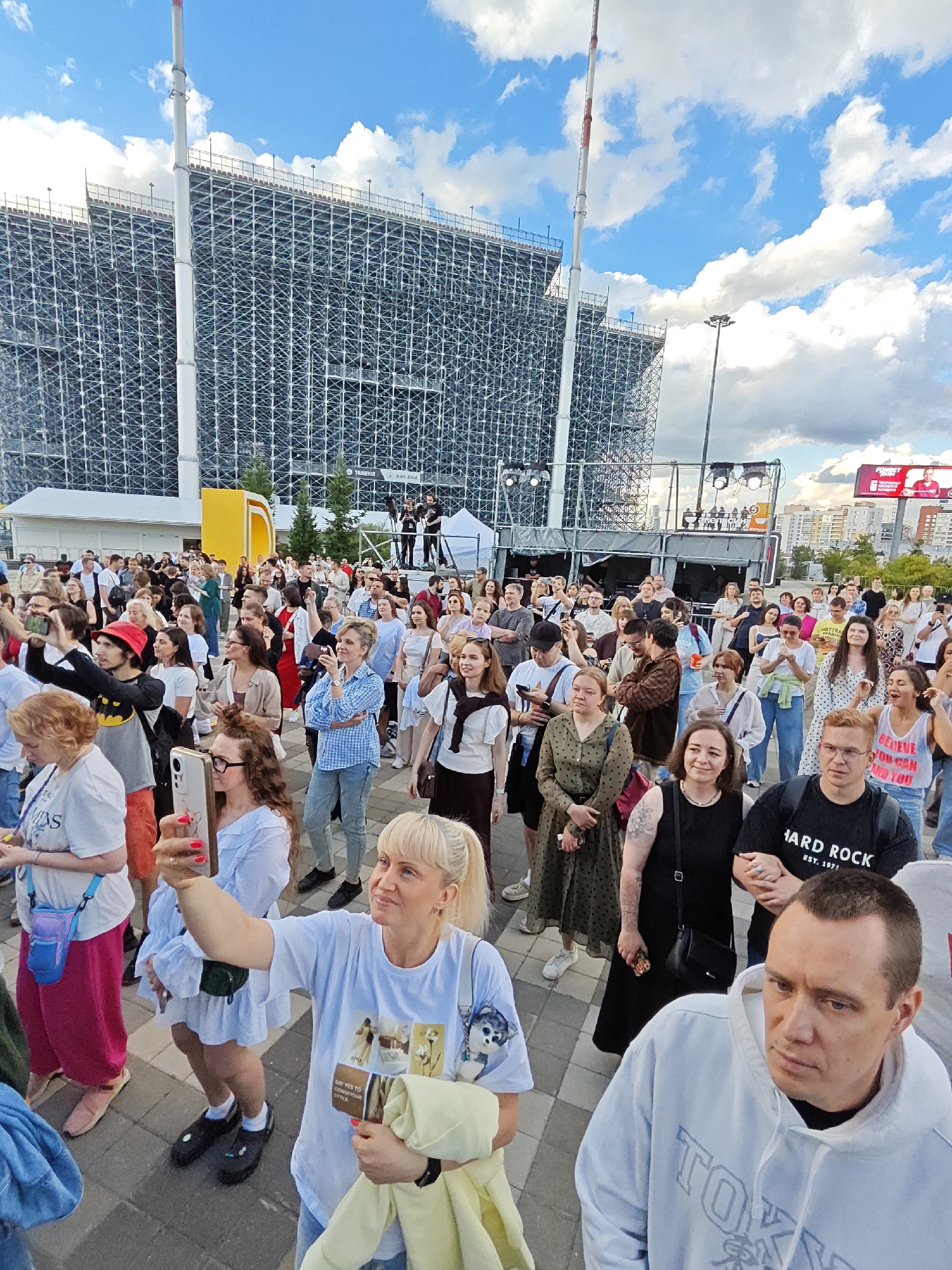 This image captures an outdoor event at the Yekaterinburg Arena in Yekaterinburg, Russia. A large crowd of people are gathered, facing a stage area that is partially obscured by a scaffolding structure. The sky above is partly cloudy with blue patches.
In the foreground, several individuals are visible. A woman in a white t-shirt is taking a selfie with her phone. To her right, a man wearing a light blue hooded sweatshirt with "TOKYO" written on it is looking away from the camera. Further to the left, a woman in a pink skirt and white t-shirt is visible.
Behind the foreground individuals, the crowd extends back towards the stage. Many people are holding up their phones, presumably to record or photograph the event. The scaffolding structure behind the stage suggests that some form of construction or renovation is taking place, or that the arena is being prepared for a large-scale event.
In the background, beyond the scaffolding, a large digital billboard displays an image of a man in red, with some text that is difficult to discern clearly. The urban environment of Yekaterinburg is visible beyond the arena, with buildings and infrastructure. The overall scene depicts a lively public gathering at an outdoor venue.