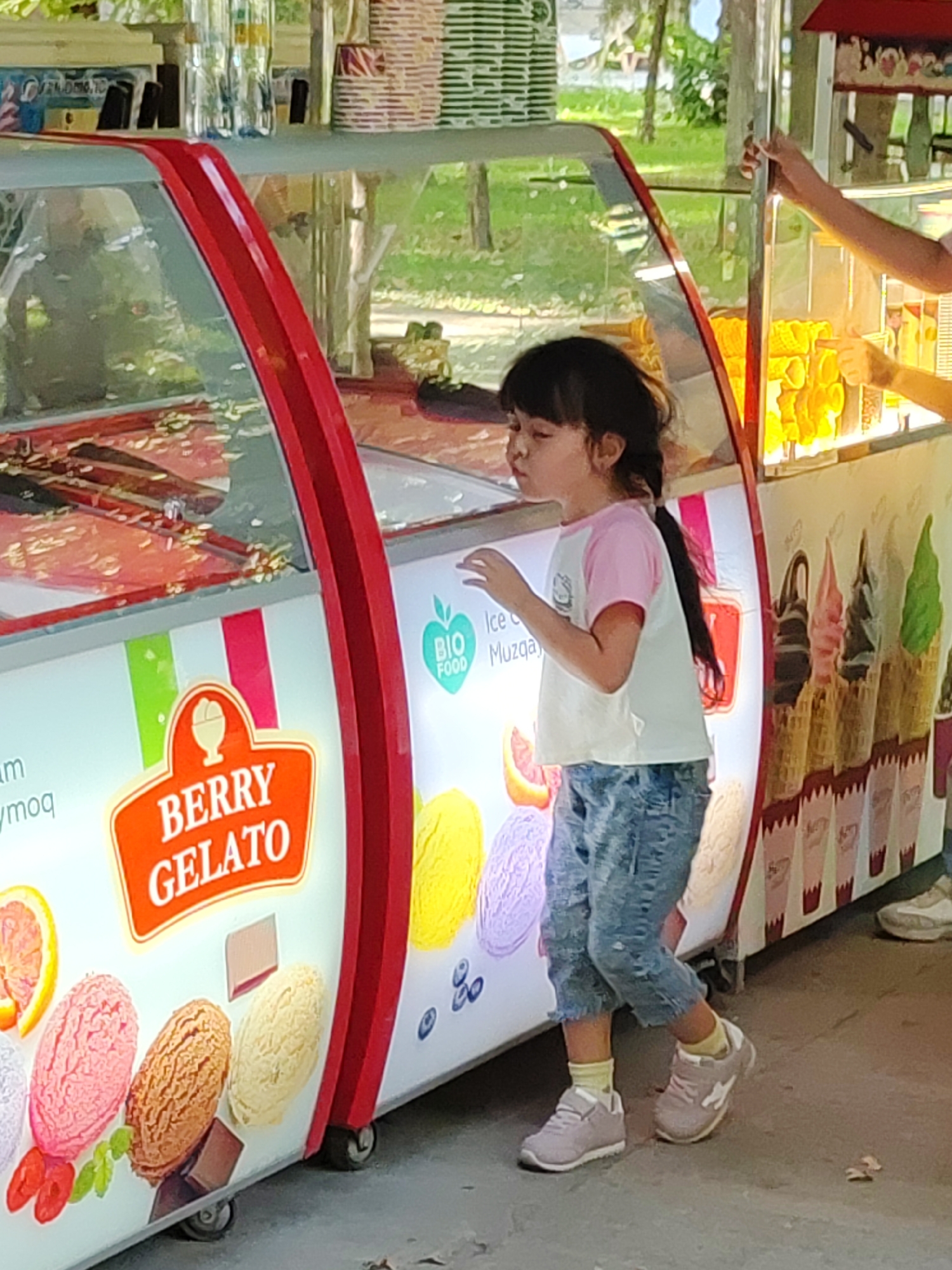 A young girl with dark hair in a ponytail, wearing a pink and white shirt and blue jeans, stands looking into an ice cream display case. The display case has red trim and a sign that reads "BERRY GELATO". Inside the case, there are various flavors of ice cream visible, including what appears to be orange, pink, brown, and yellow. To the right of the display case, another person's hand is reaching into the case, possibly serving ice cream. The background suggests an outdoor setting with greenery and trees. The image was taken in Tashkent, Uzbekistan.