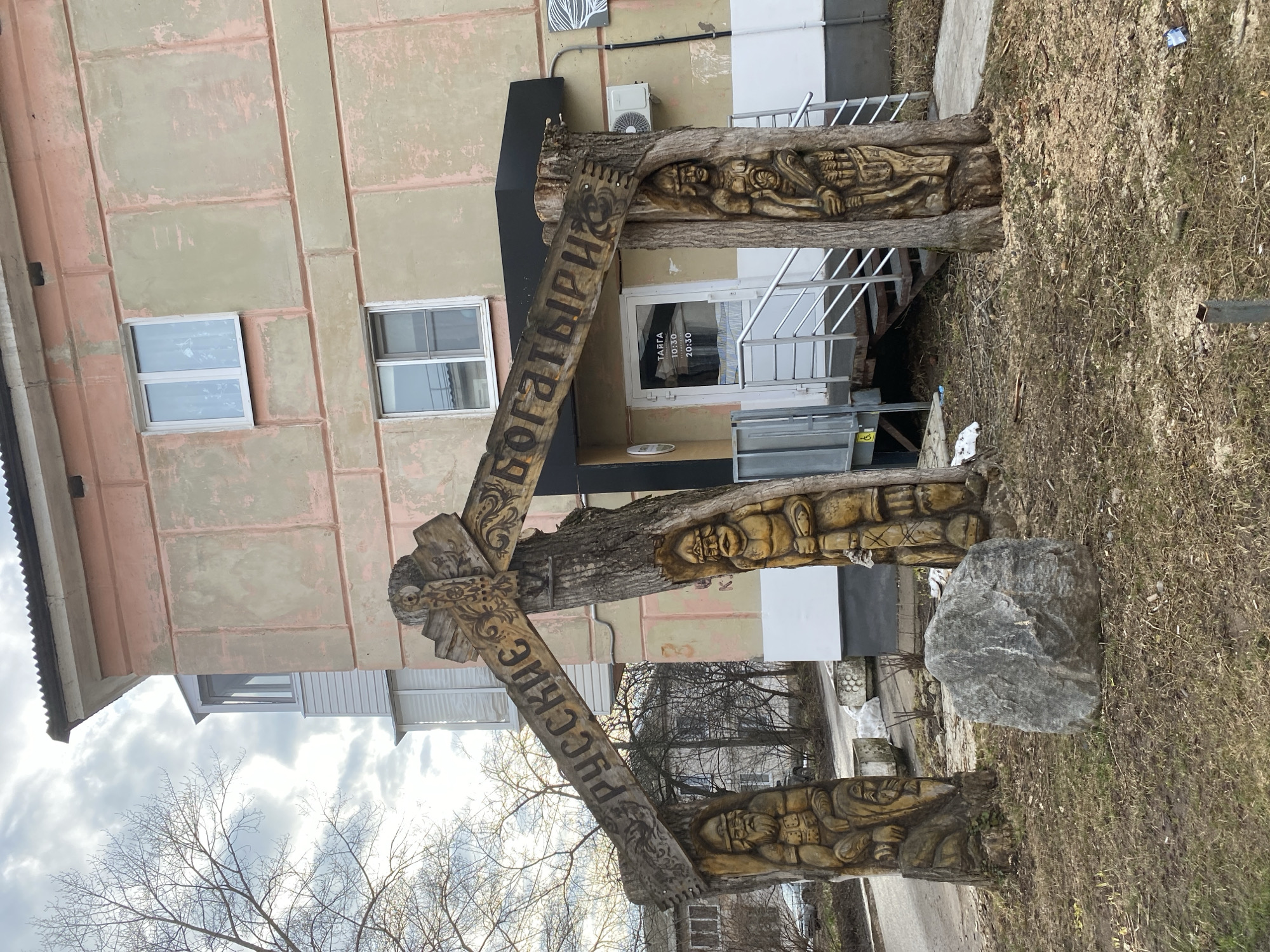 The image shows a wooden sculpture in the city of Chaikovsky, Russia. The sculpture is composed of three upright tree trunks with carvings of figures, connected by two diagonal beams. The beams are inscribed with the Cyrillic words "РУССКИЕ БОГАТЫРИ" (Russian Bogatyrs). The sculpture is located in front of a building with two windows and a white door, accessible by a set of stairs. A large grey rock sits near the base of one of the tree trunks. The ground is covered in dry grass and debris. A bare tree is visible in the background against a cloudy sky.