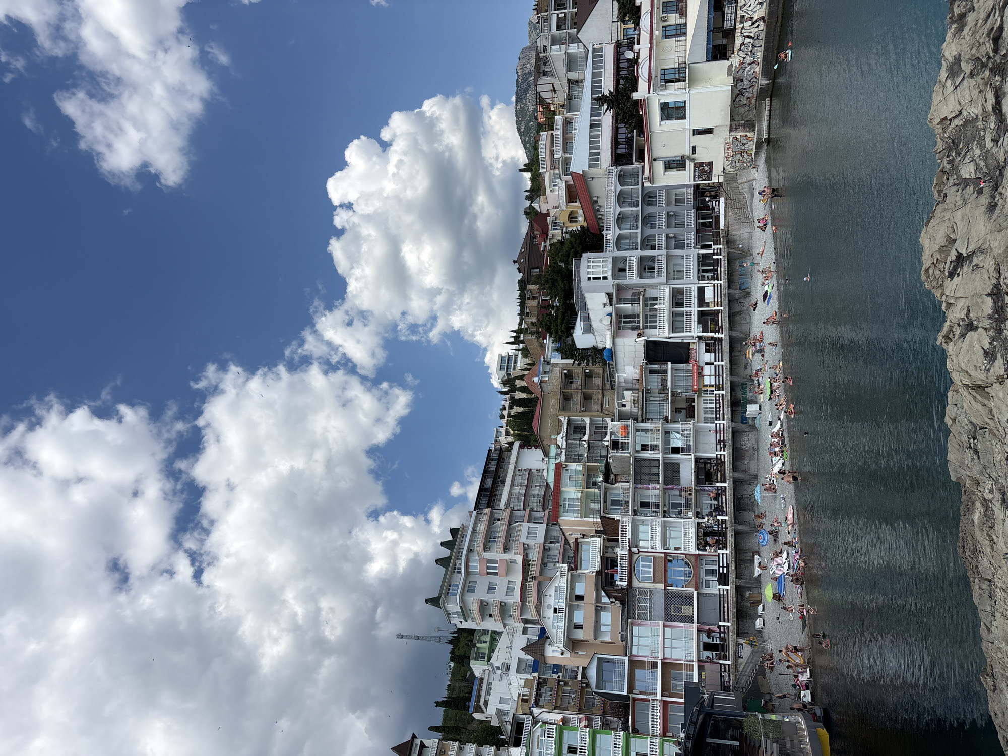 The image depicts a coastal town with multi-story buildings constructed on a hillside overlooking the sea. The buildings are a mix of modern and older architecture, with many featuring balconies and colorful facades. A beach is situated at the base of the buildings, populated by numerous individuals sunbathing and swimming. The sea is calm and dark blue. The sky above is partly cloudy with fluffy white clouds against a bright blue background. The scene appears to be a popular tourist destination, likely during the summer season. The location is identified as Kiparisnoe, Ukraine.