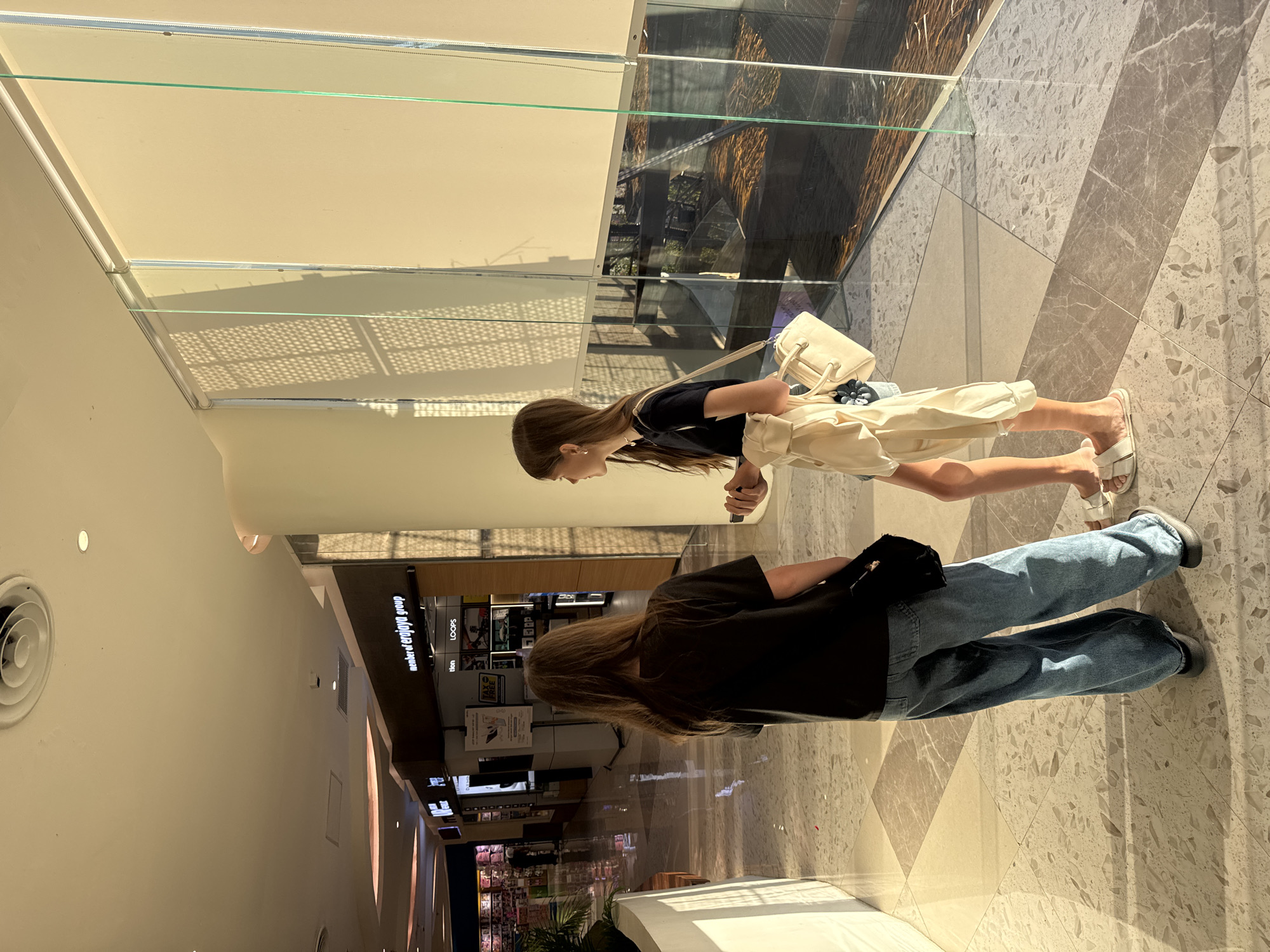 Two young women are captured in a bright, modern indoor setting, likely a shopping mall in Denpasar, Indonesia. The image is oriented vertically, showing the subjects and surroundings from an overhead perspective if viewed horizontally.
One woman, positioned closer to the foreground and seen from the side, has long, light brown hair. She wears a black short-sleeved top, light cream-colored wide-leg pants or a skirt, and white open-toed sandals. She carries a small, light-colored handbag with a top handle and a shoulder strap. Her gaze is directed forward.
The second woman is seen from behind, positioned further back and to the left. She has long, dark brown hair and is dressed in a black t-shirt and blue jeans. A dark item, possibly a bag, is tucked into the back of her waistband.
The environment is spacious and well-lit, with strong natural light entering from above, creating distinct shadows on the floor. The floor itself is made of large, light-toned tiles with a speckled or terrazzo-like pattern, arranged in geometric shapes. Glass railings or barriers are prominent, suggesting multiple levels or architectural features within the building. In the background, various shop fronts and display cases are visible.
Visible text on store signage includes "member of erajaya group," "LOOPS," and partially obscured words like "tlon," "MAX," and "ATM." The scene suggests a typical daytime mall visit, with the individuals either walking or pausing to observe their surroundings.
