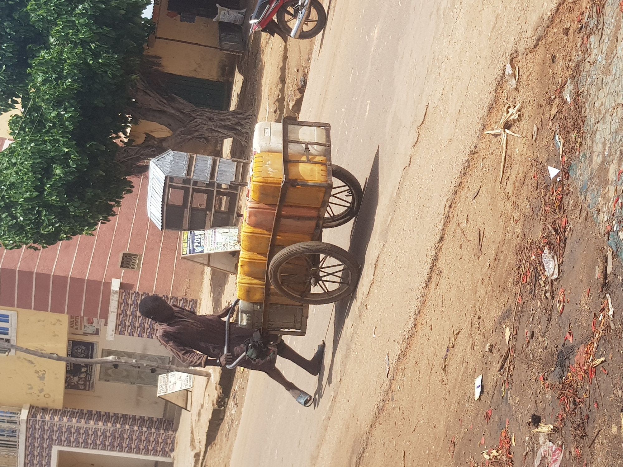 In Tukur Tukur, Nigeria, a dark-skinned individual wearing a brown garment is pushing a cart loaded with yellow and white plastic containers filled with liquid. The cart has two spoked wheels. The individual is walking on a paved road with a dusty shoulder. In the background, there are buildings, including one with a yellow facade and another with red brick. A large tree with green foliage stands behind the cart. A red motorcycle is parked on the side of the road. Debris, including plastic and organic matter, is scattered on the ground.