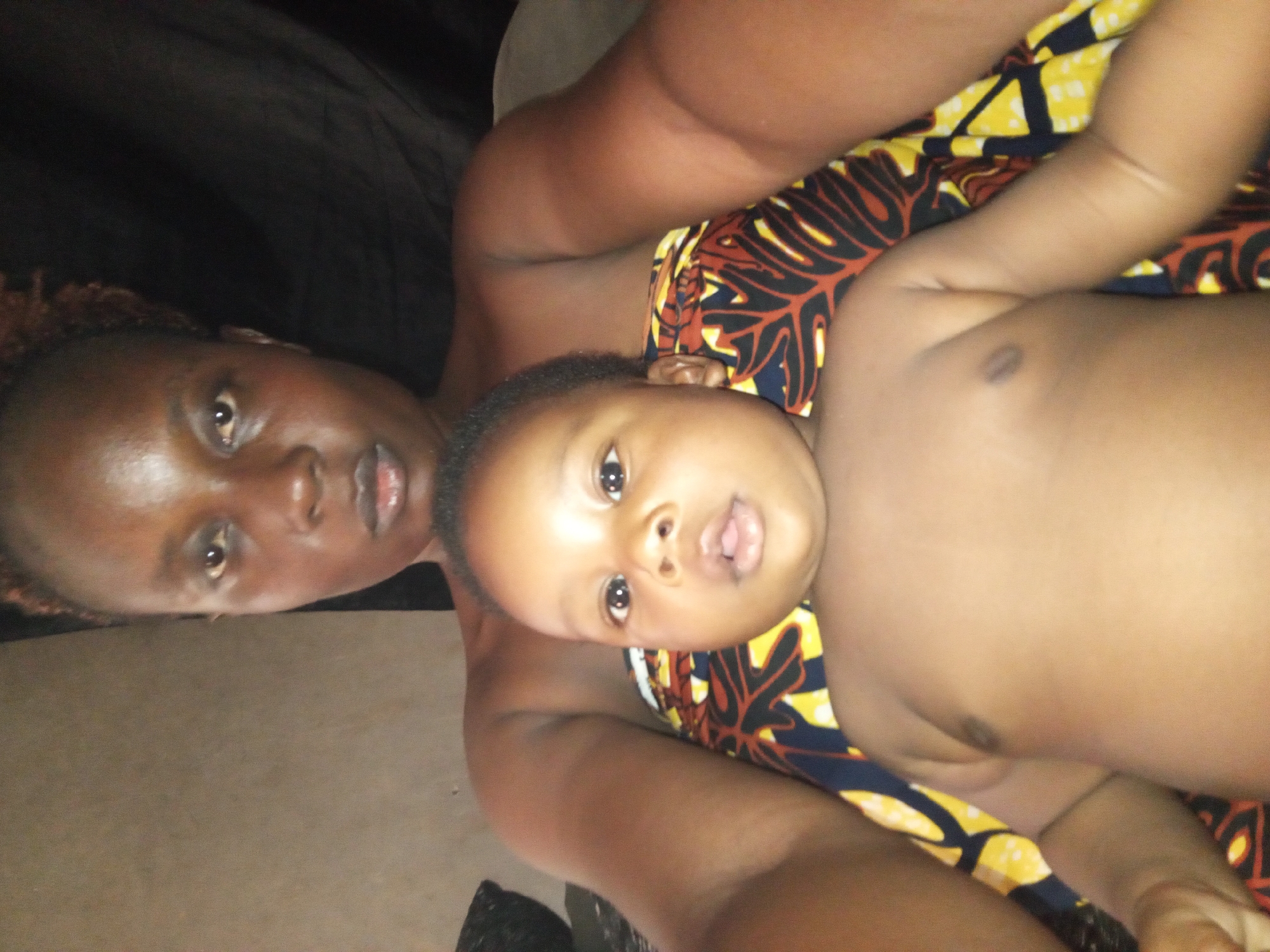 An adult and a baby are featured in a close-up, rotated image. The adult, with dark skin and short, reddish-brown hair, is positioned on the left side of the frame, looking directly towards the viewer with a neutral expression. Only their head, shoulder, and arm are fully visible, with a dark fabric behind them.
The baby, also with dark skin and short, dark hair, lies to the right of the adult, with their head resting near the adult's arm. The baby has wide, dark eyes and a slightly open mouth, looking towards the camera with an expression of curiosity or wonder. A vibrant, colorful fabric, possibly an African print with yellow, red, and black patterns, is draped around the baby's body. The adult's arm is visible under the baby's head, and part of the baby's torso is uncovered.
The setting appears to be indoors, with a light brown/beige surface visible in the background behind the adult and beneath the baby. The lighting is somewhat dim, suggesting an intimate, indoor moment. The location context provided is Sabon Tasha, Nigeria. There is no visible text in the image.